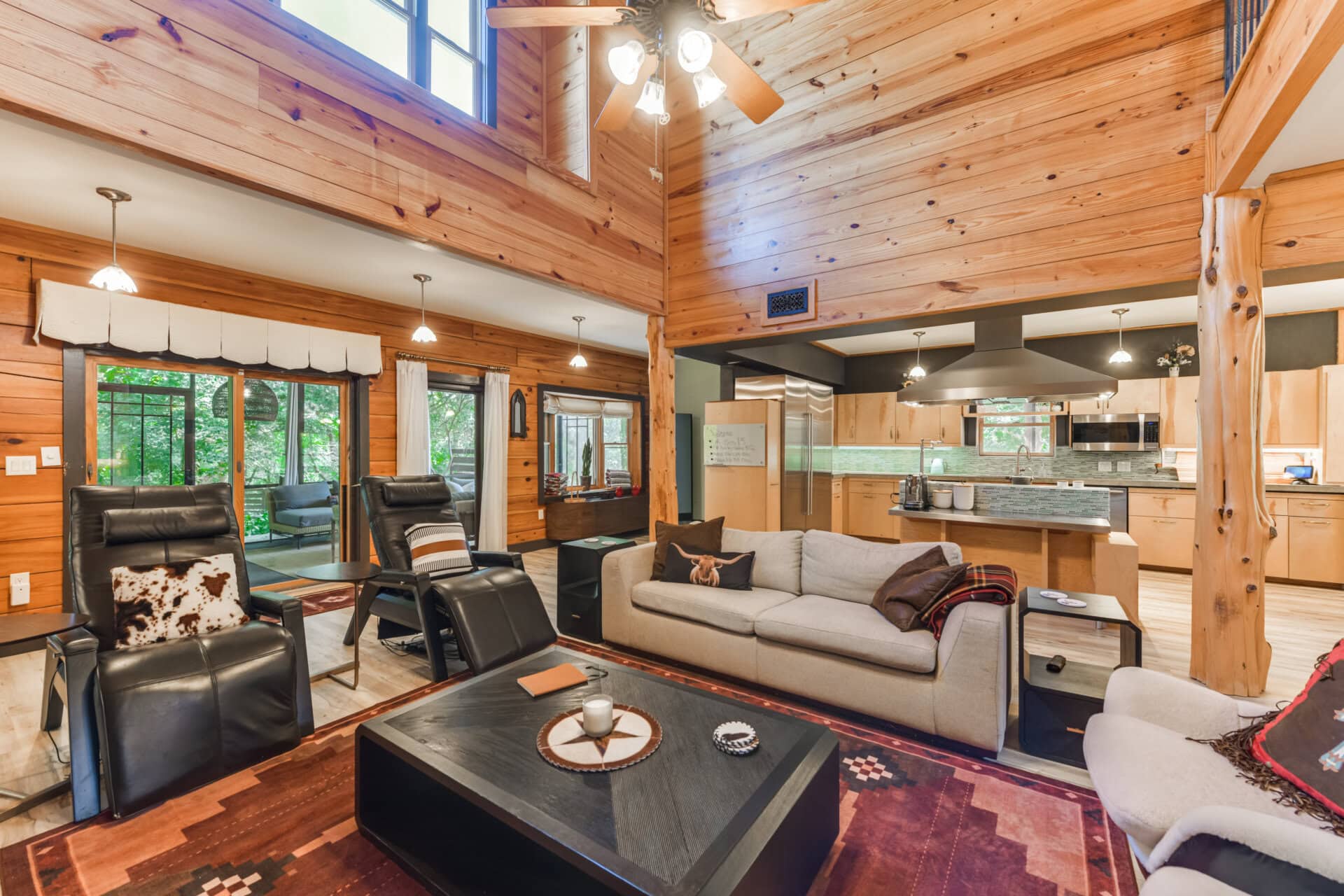Cozy cabin living room with wood walls and vaulted ceiling, leather recliners, a gray sofa, and an open kitchen in the background.