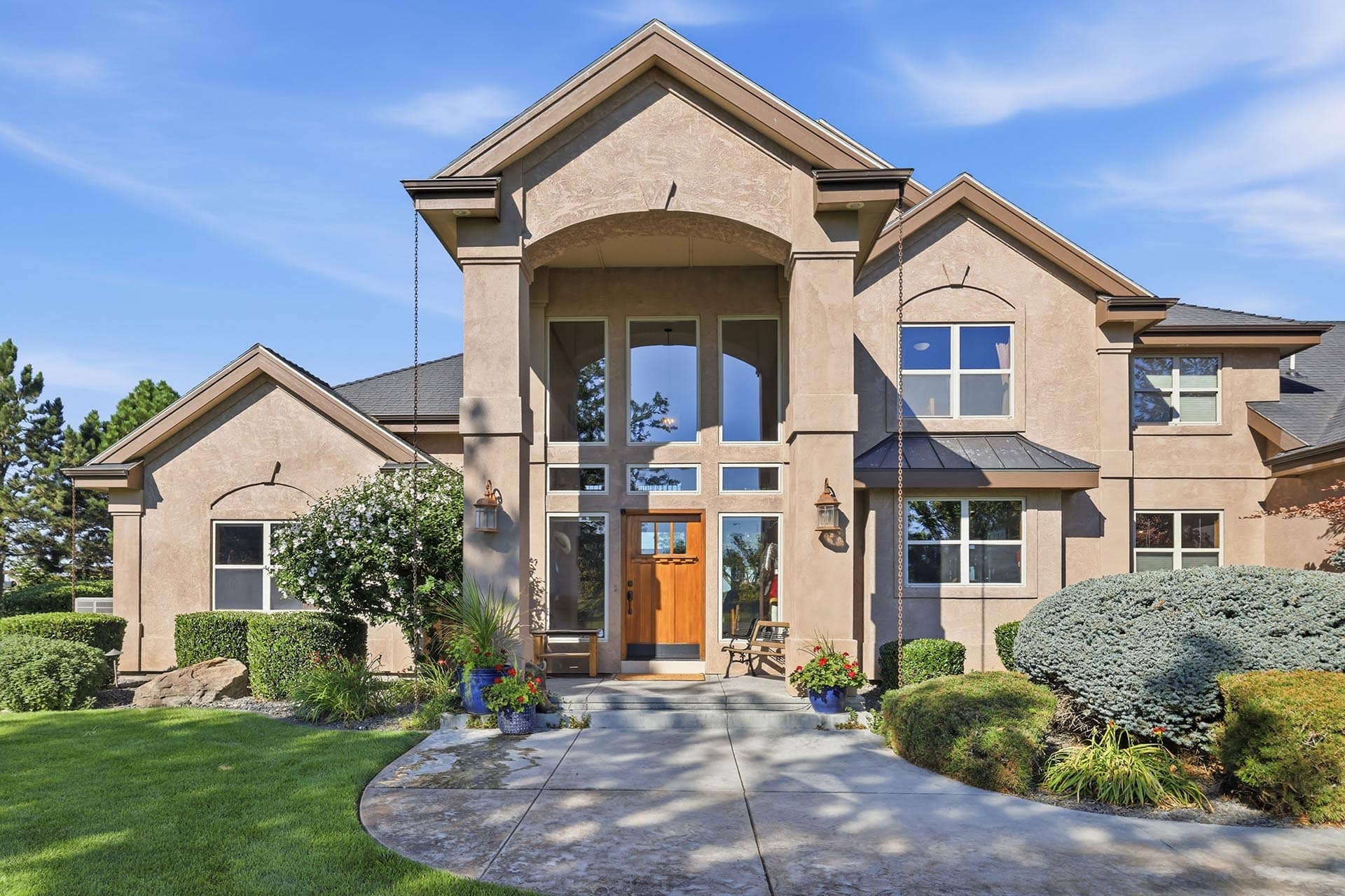 Front exterior of a beige stucco house with a wooden front door, large windows, and a well-kept lawn and shrubs under a clear blue sky.
