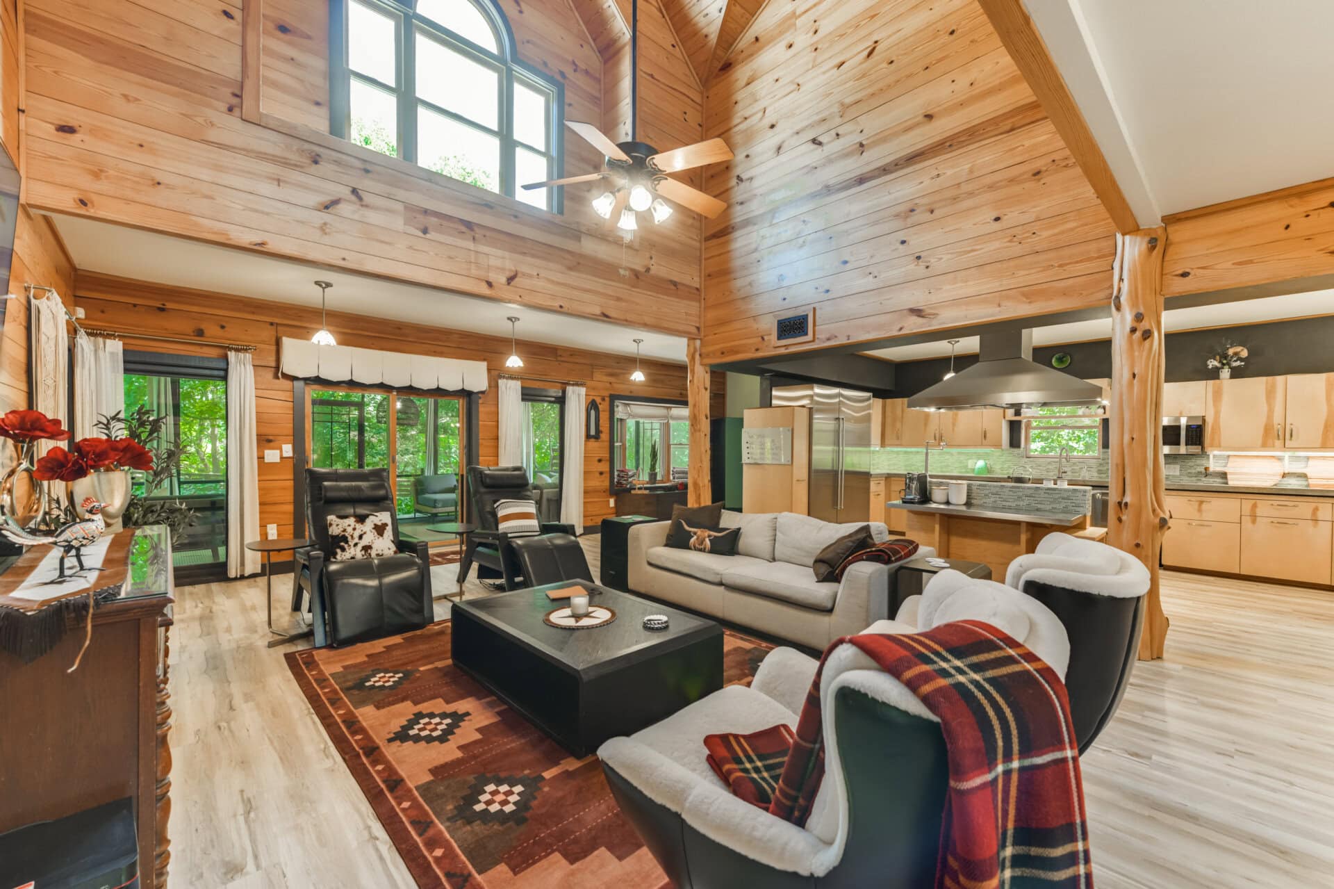 Open living area with vaulted wood ceiling, large windows, and a modern kitchen; dark leather chairs around a black coffee table.