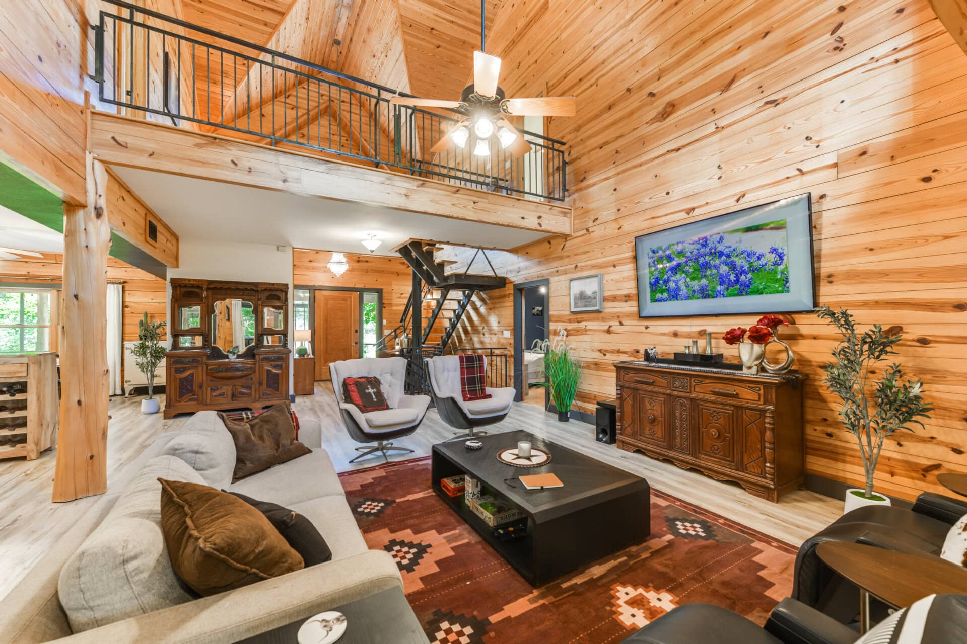Cozy wood-paneled living room with a loft, staircase, and burgundy rug; beige sofa and vintage wooden furniture.