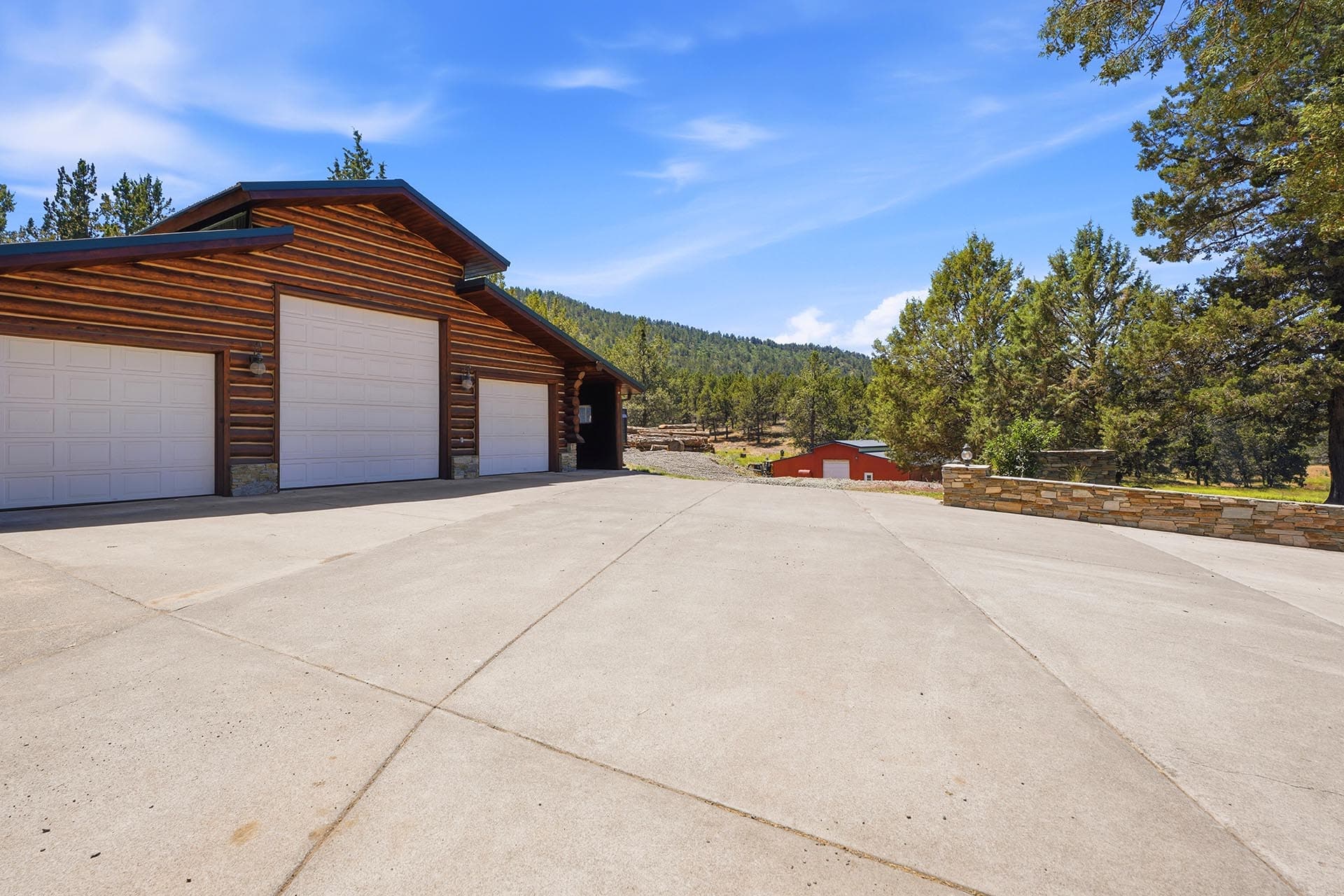 Log cabin-style garage with three white doors, stone bases, and a wide concrete driveway set against a forested backdrop and blue sky.