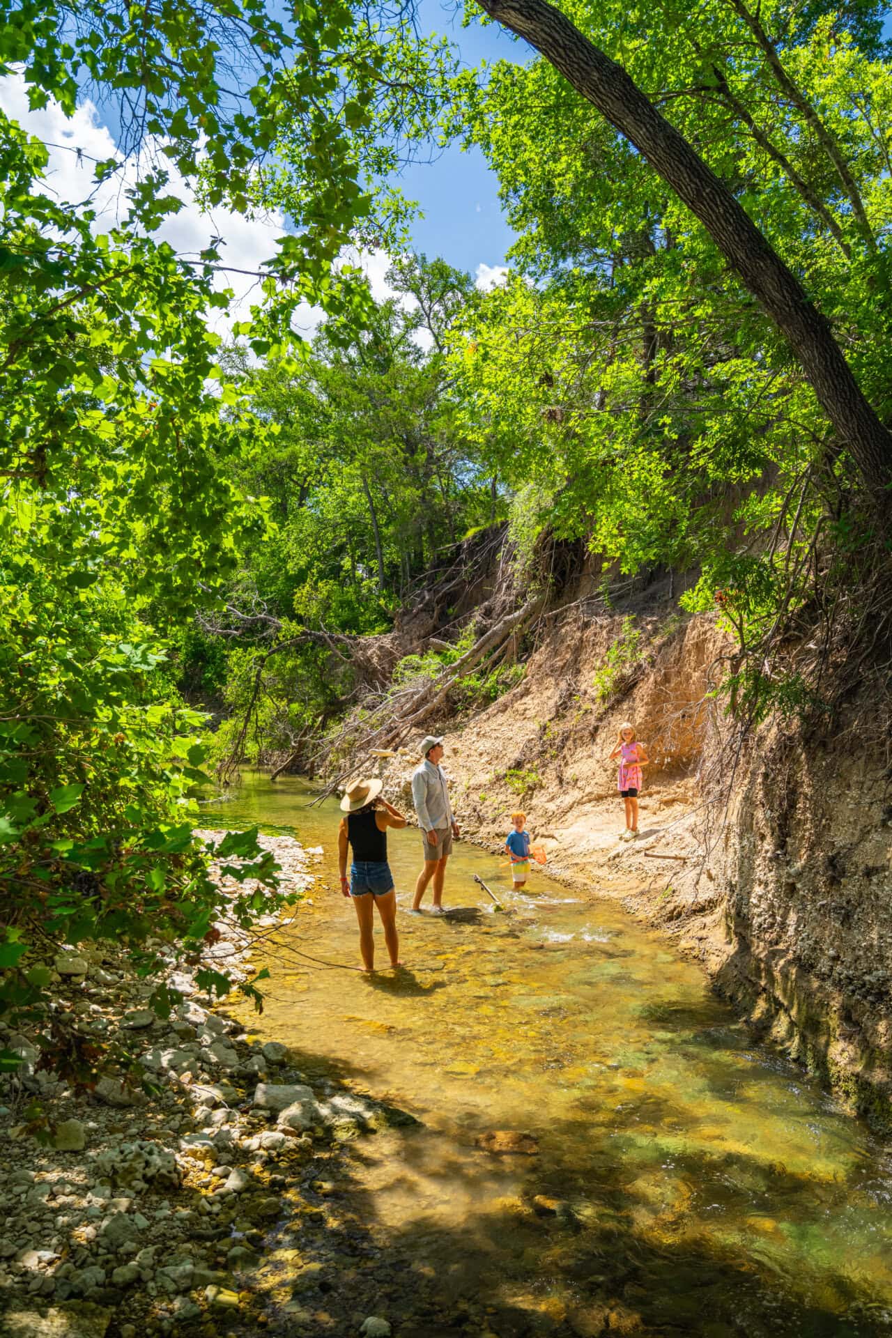 Family wading in a shallow, clear river along a rocky, tree-lined bank on a sunny day.