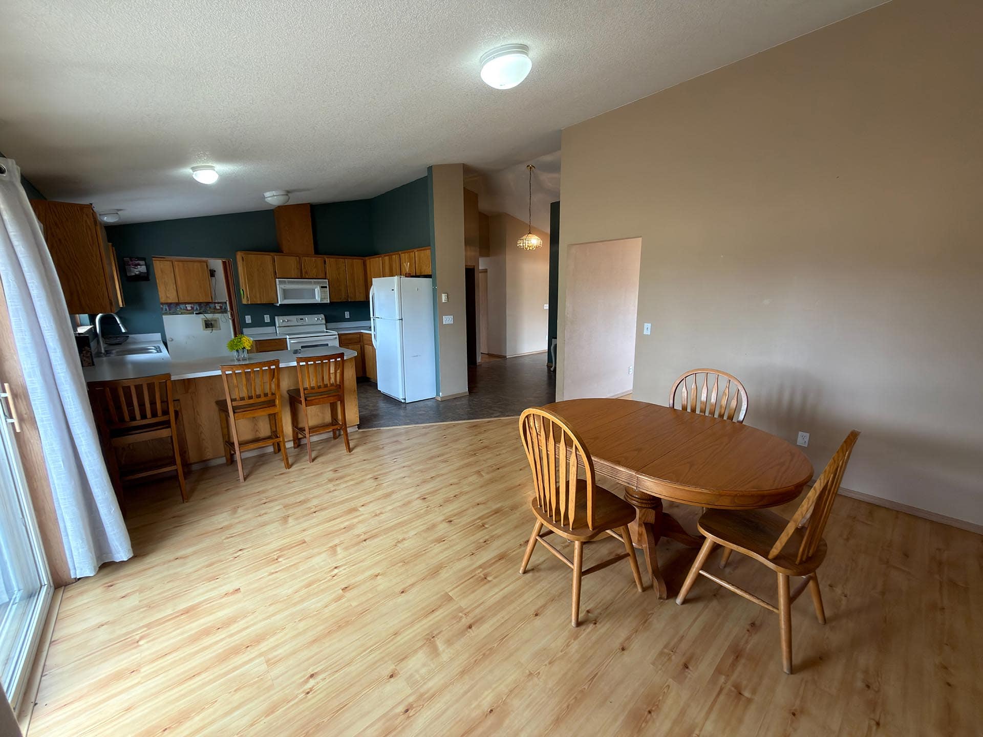 Open-plan kitchen and dining area with wooden cabinets, white appliances, and a round wooden dining table with chairs near a sliding door with curtains (light wood floor).
