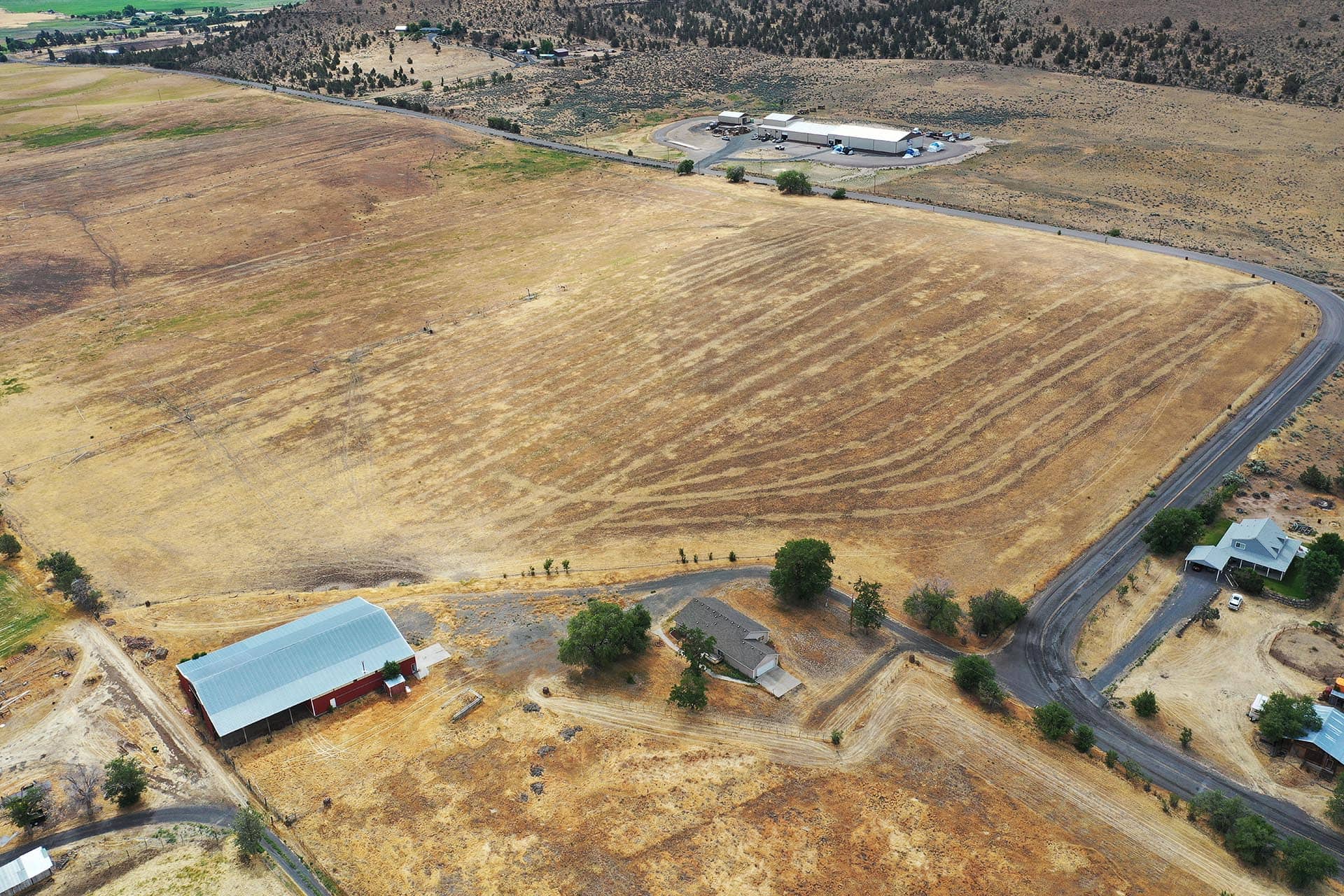 Aerial view of a dry, brown farmland with a curved road and a red barn near the bottom left, surrounded by scattered trees and houses.