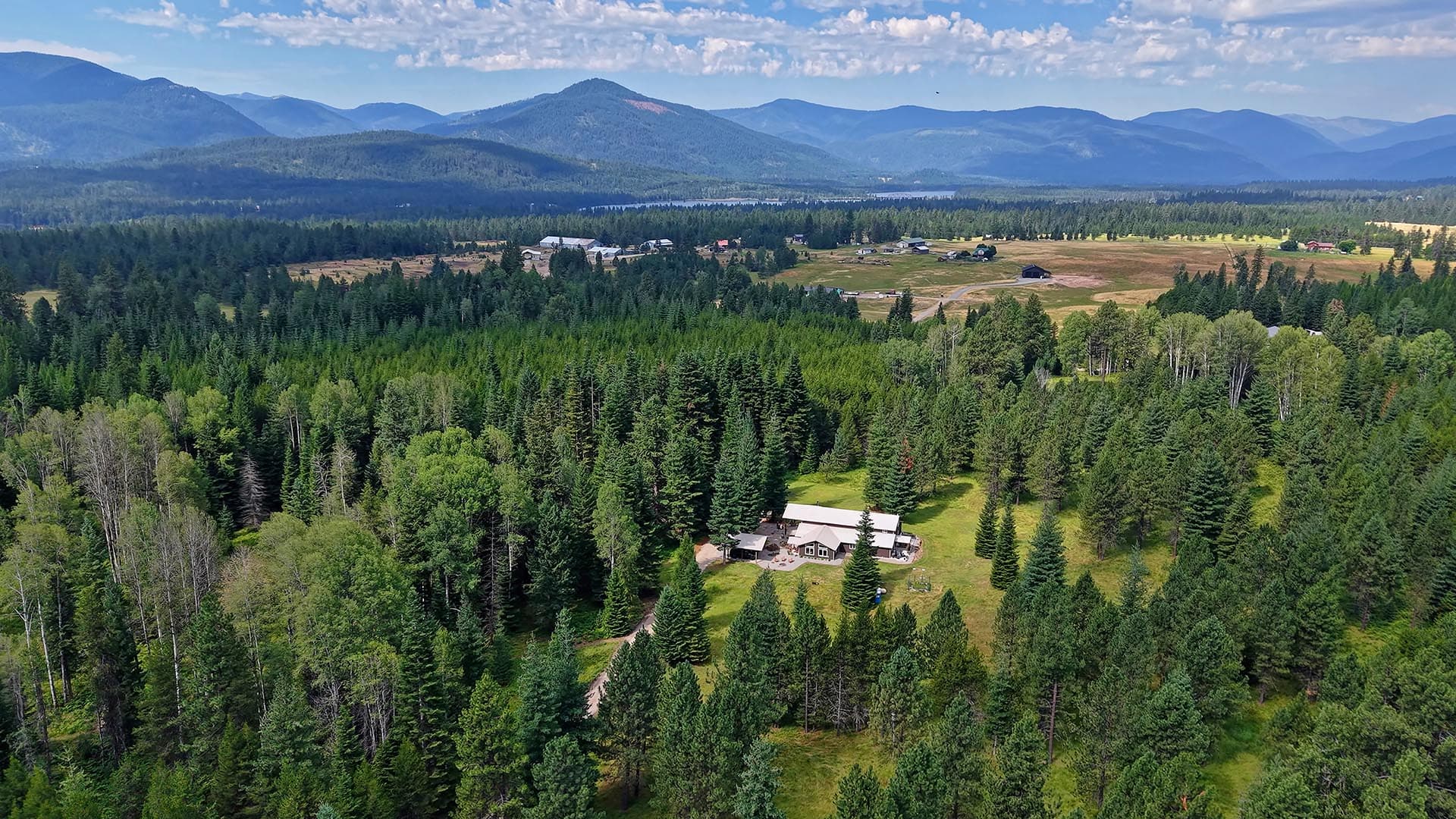Aerial view of a forested valley with a few farm buildings and distant blue mountains under a partly cloudy sky.