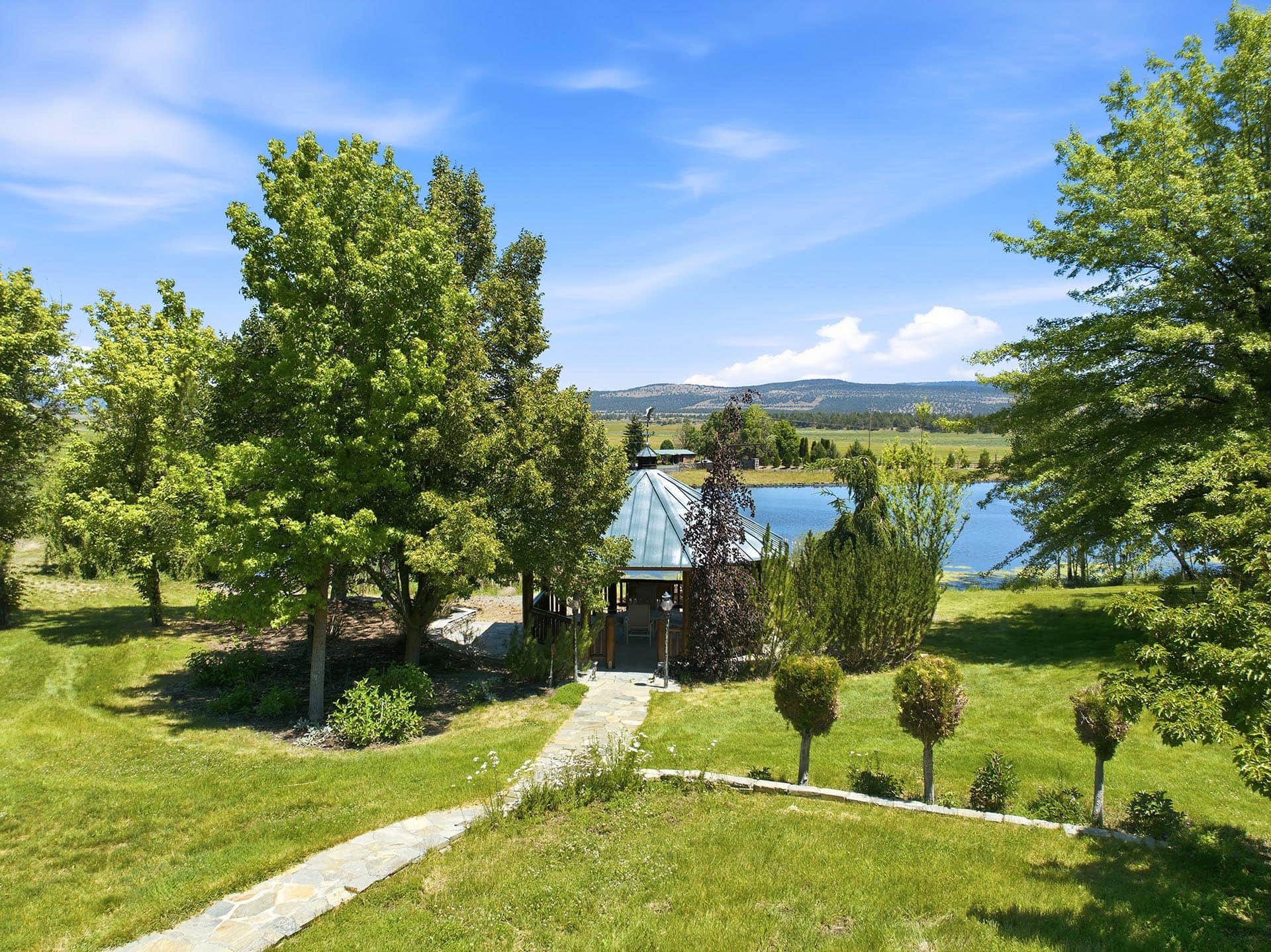 Stone path through a green garden leads to a gazebo by a lake, with trees and hills beyond.