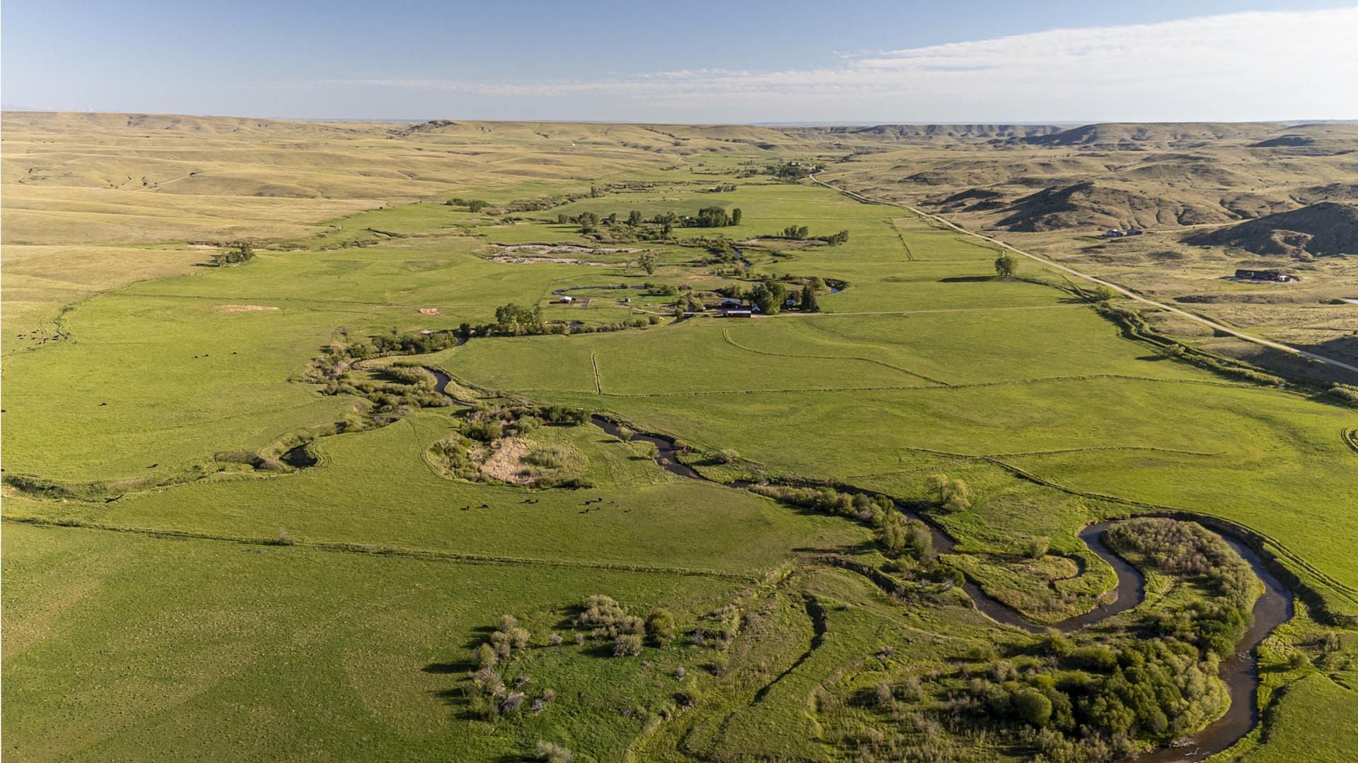 Aerial view of green farmland with a winding river, scattered trees, and farm buildings set against rolling brown hills in the distance.