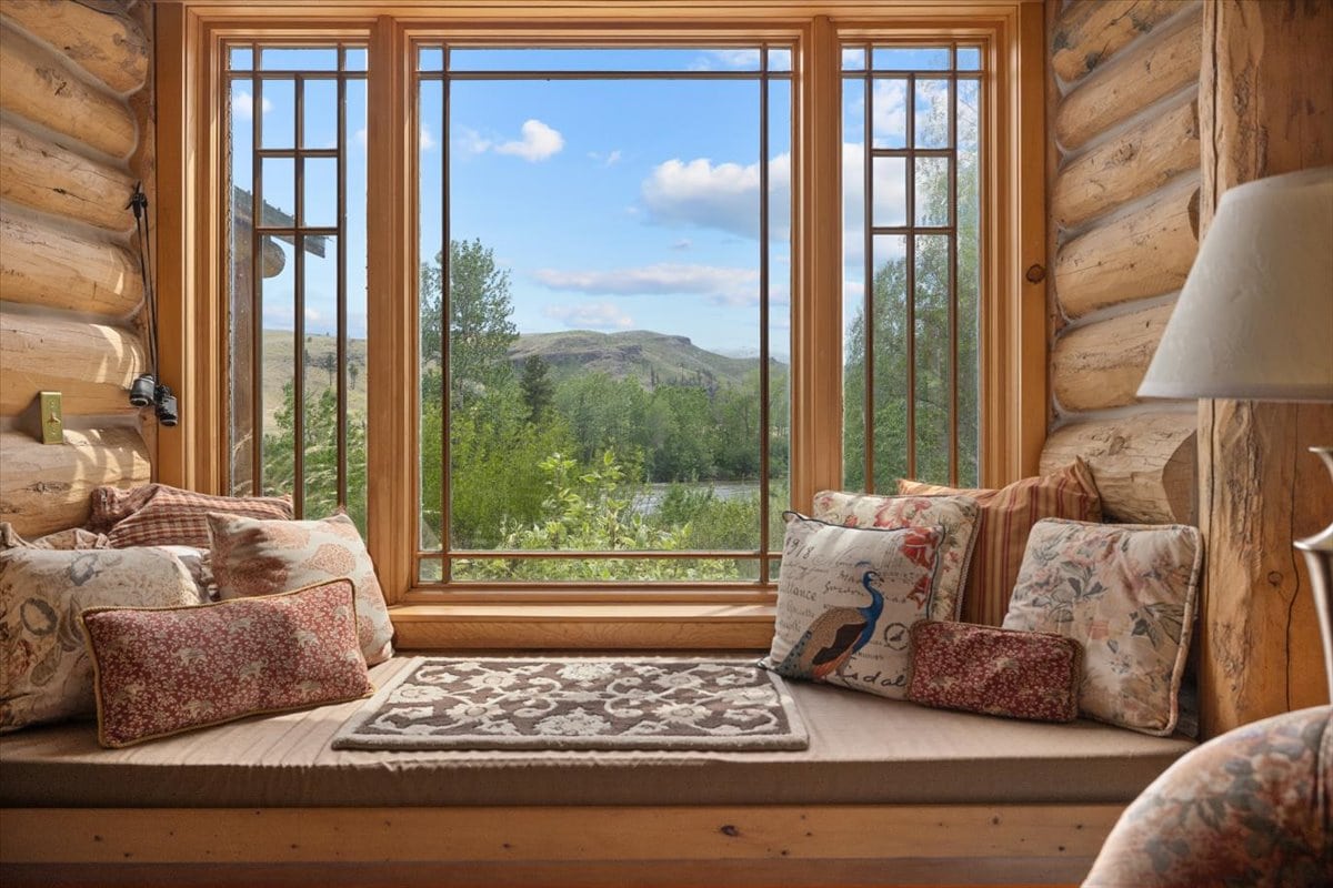 Cozy log-cabin window seat with patterned cushions, overlooking trees and distant blue hills under a bright sky.