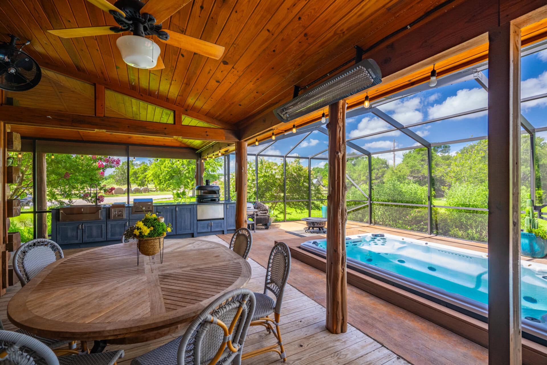 Wooden-paneled outdoor patio with a round table and wicker chairs, ceiling fans, and string lights overlooking a fenced yard and hot tub area.