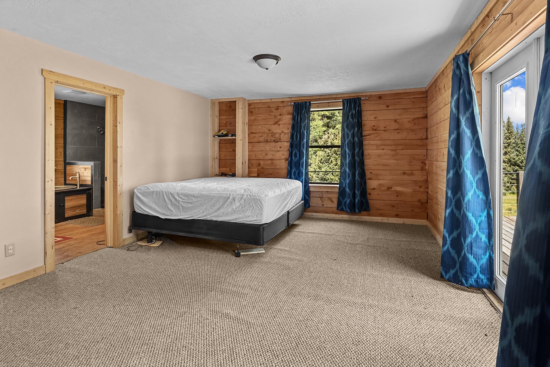 Bedroom with wood-paneled walls, a raised bed on a platform, and blue patterned curtains; doorway to a bathroom on the left and a window toward trees outside.