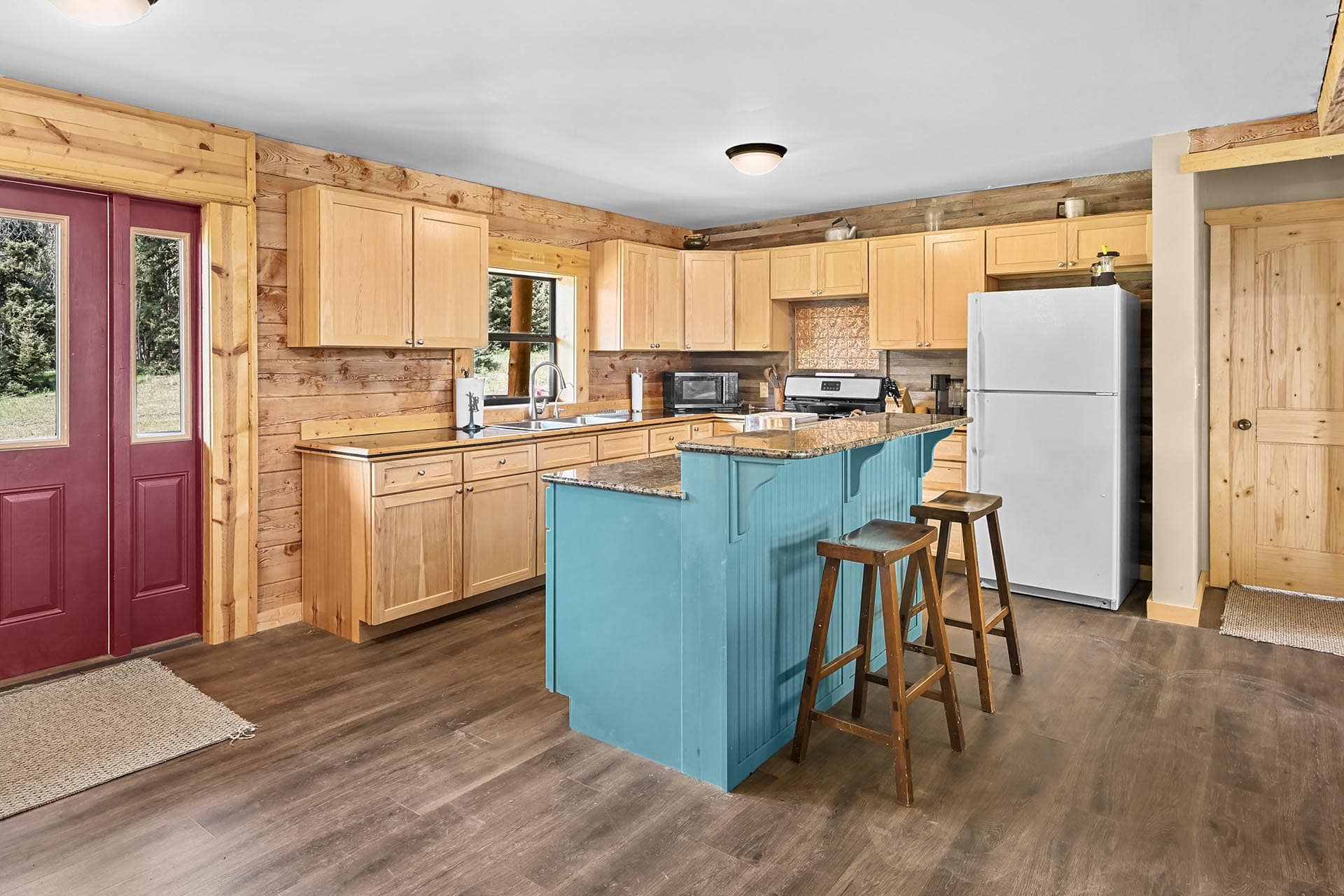 Rustic kitchen with wood walls, light wood cabinets, a turquoise island, stainless fridge, and a red front door visible on the left.