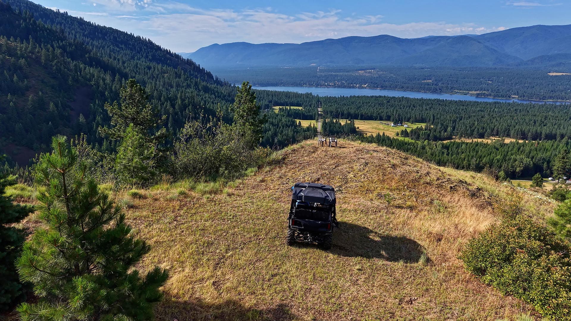 Off-road vehicle parked on a grassy overlook as pine trees frame a valley, lake, and distant mountains under a blue sky.