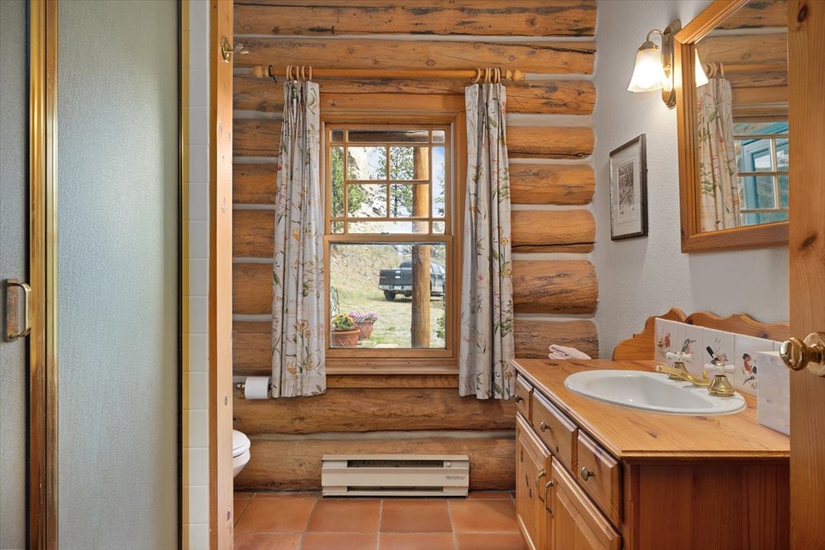 Rustic log-cabin bathroom with a wooden vanity, oval sink, brass faucet, and a window with floral curtains; outdoor view shows a truck.