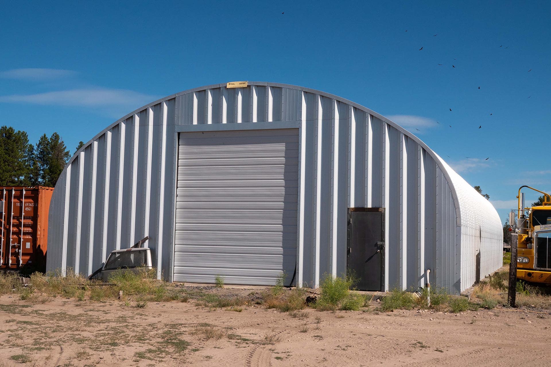 Metal arched warehouse with a large roll-up door in a dusty yard, with a yellow construction vehicle on the right and a shipping container on the left. Visual: clear blue sky with birds above.