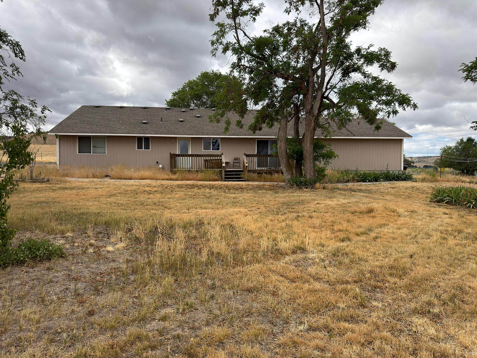 Single-story beige house with a wooden deck, set in a dry, grassy yard under a cloudy sky, with a large tree near the deck.