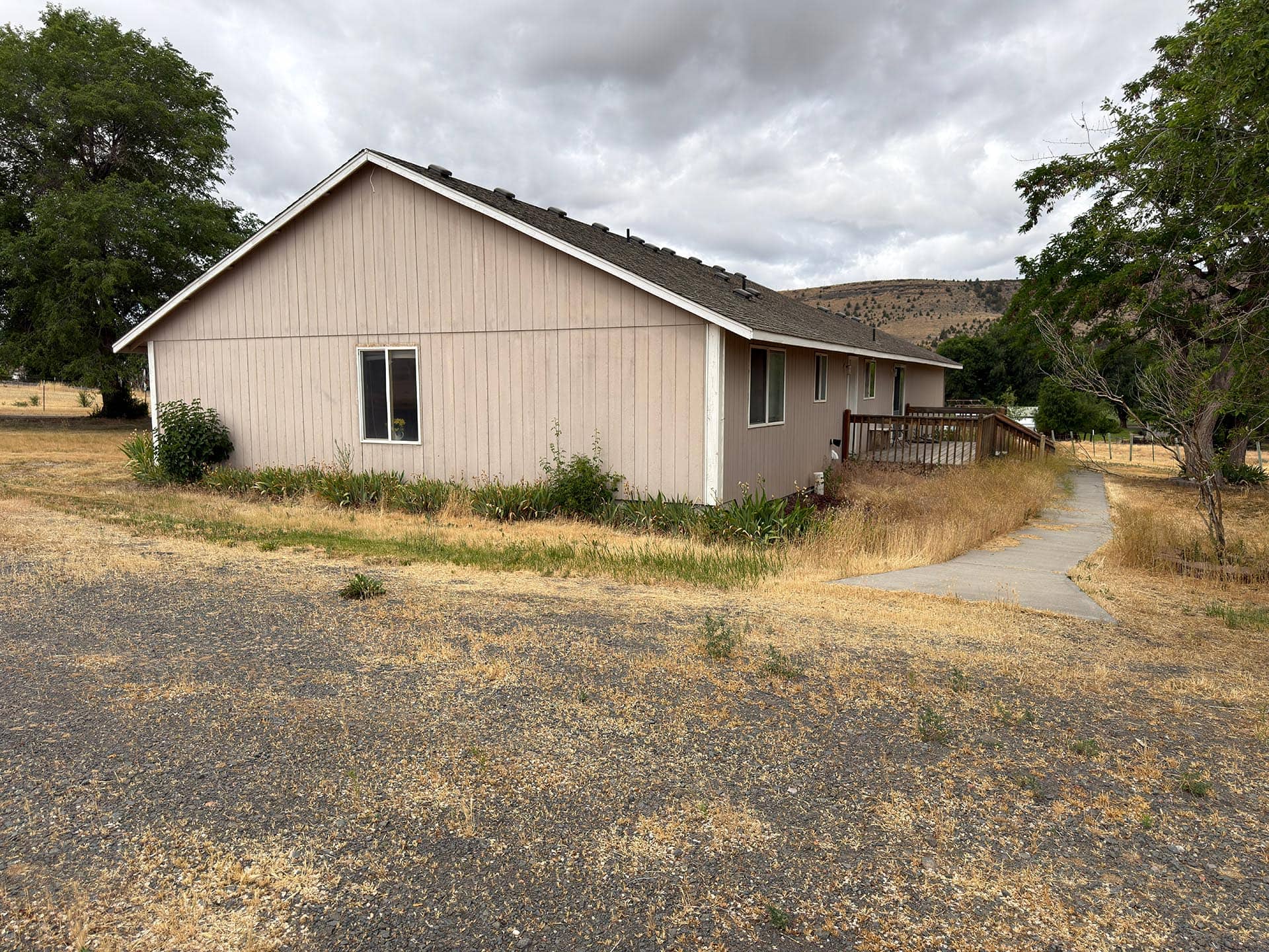 Beige single-story house with a sloped roof and wooden deck, set in a dry grassy yard with cloudy sky above.