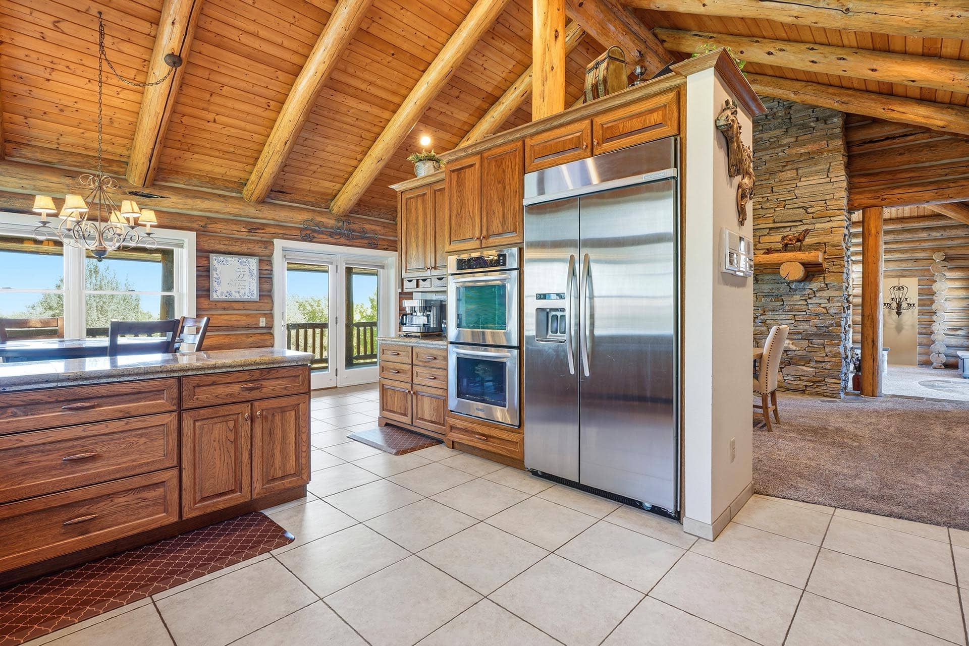 Open-concept kitchen in a log cabin with a stainless steel french-door refrigerator, double ovens, and wooden cabinets under a vaulted log ceiling.