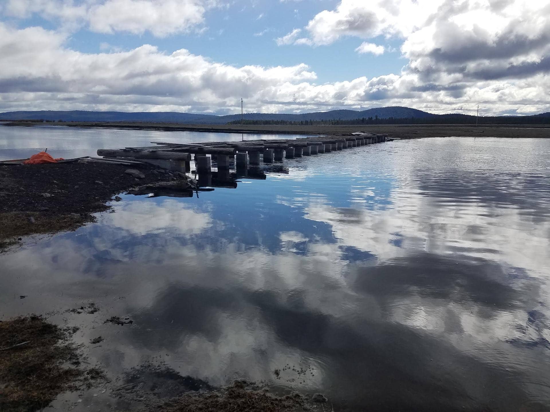 Wooden pier extends into a calm lake, reflecting cloudy skies and distant hills on a gray day.