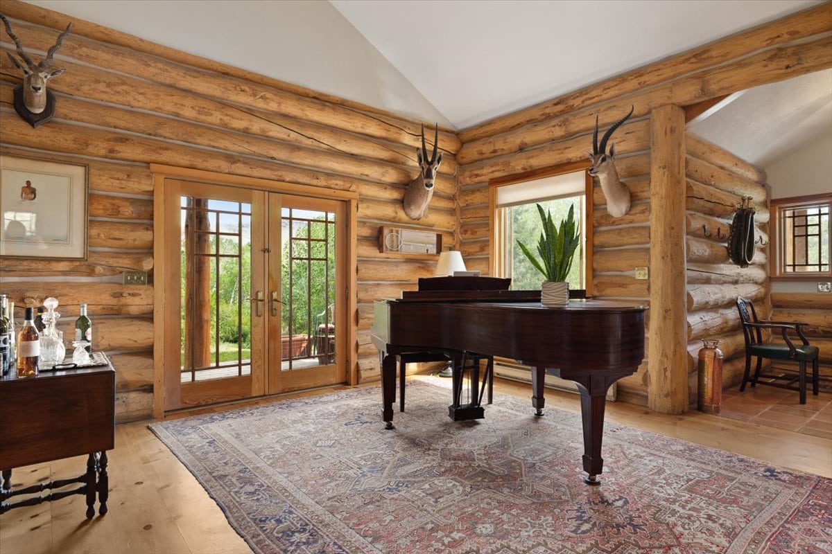 Rustic living room with a dark grand piano on a patterned rug, log walls, and French doors leading outside.