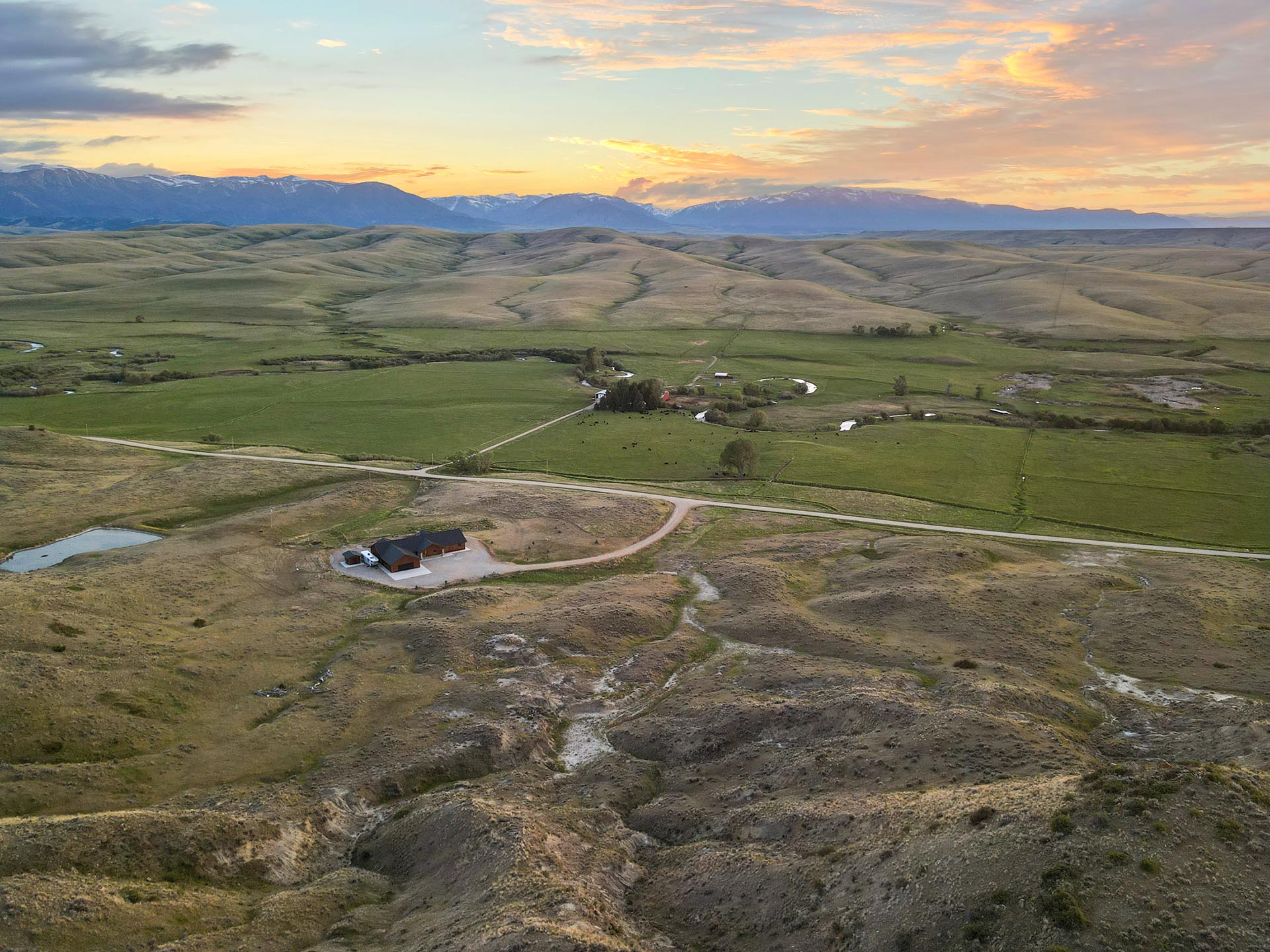 Aerial view of a rural valley at sunset with green fields, a winding river, scattered farm buildings, and distant mountains behind.