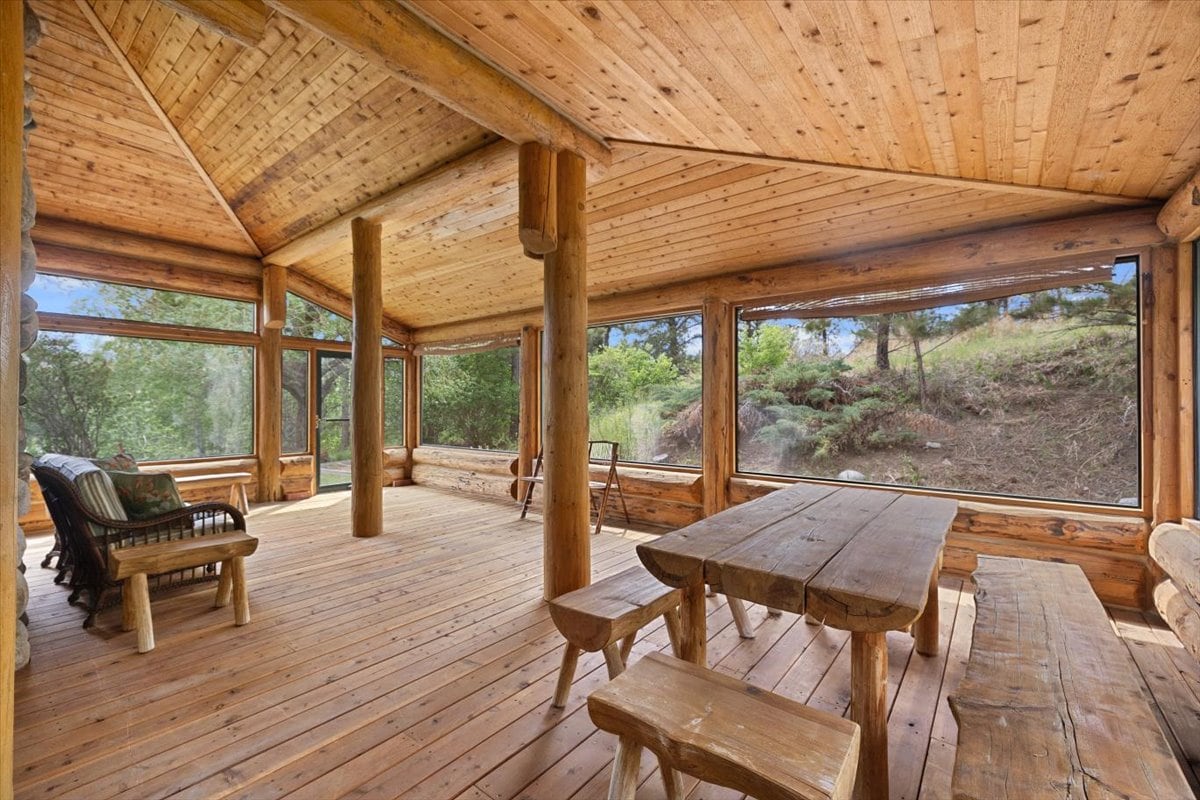 Rustic log cabin sunroom with a large wooden table and benches, log beams, and expansive windows looking out onto a forested hillside.