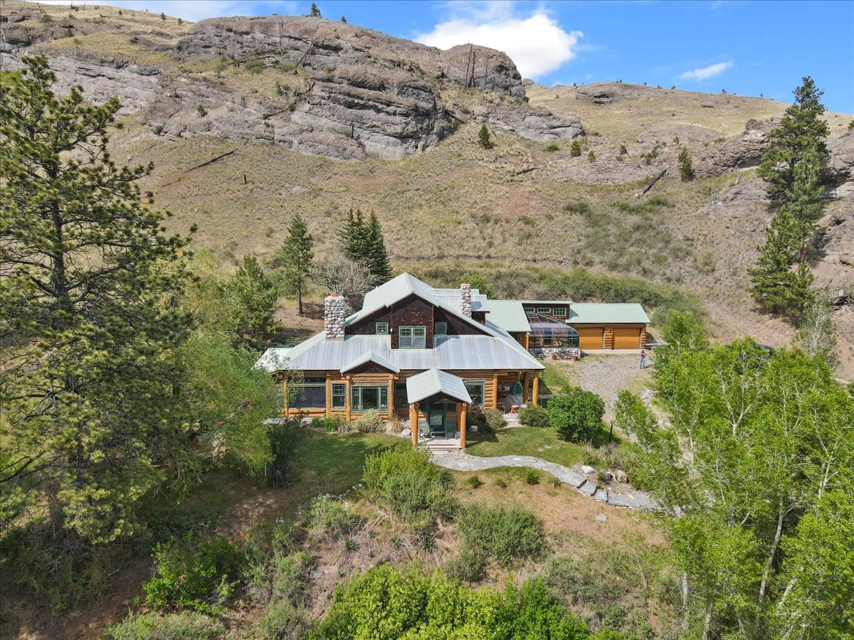 Rustic log cabin with metal roof and stone chimneys on a rocky hillside, surrounded by trees; a detached garage is to the right.