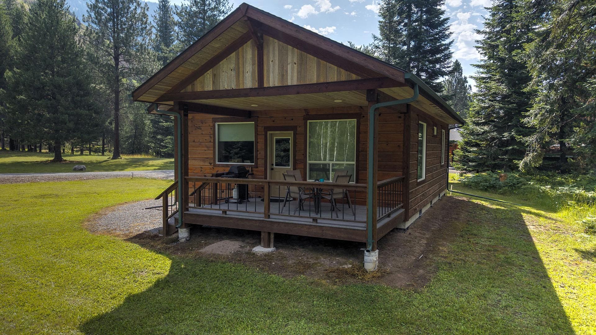 Wood cabin with a covered front porch, outdoor table and chairs, in a grassy, tree-filled yard.