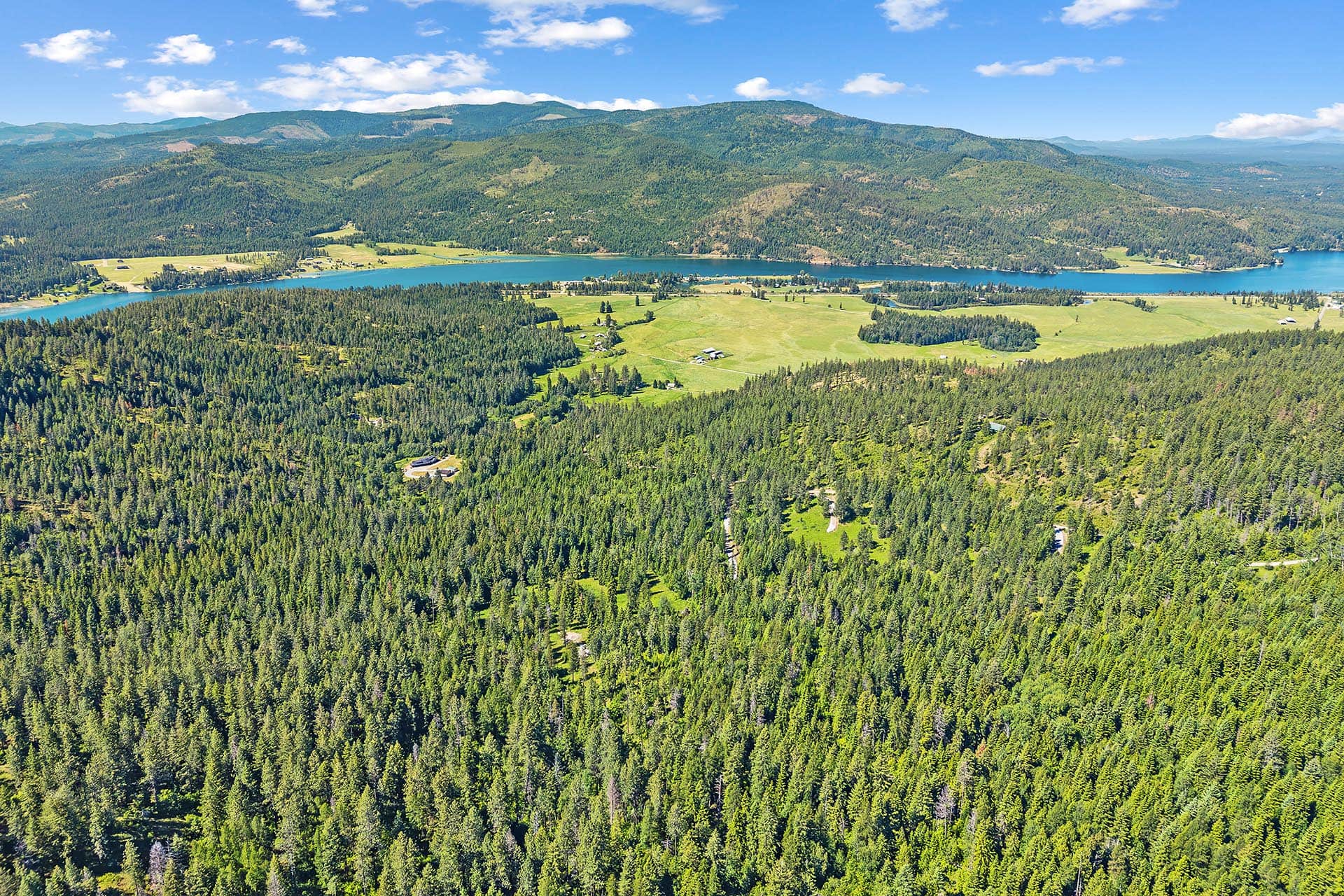 Aerial view of a forested valley with a turquoise lake and green fields beneath a bright blue sky.
