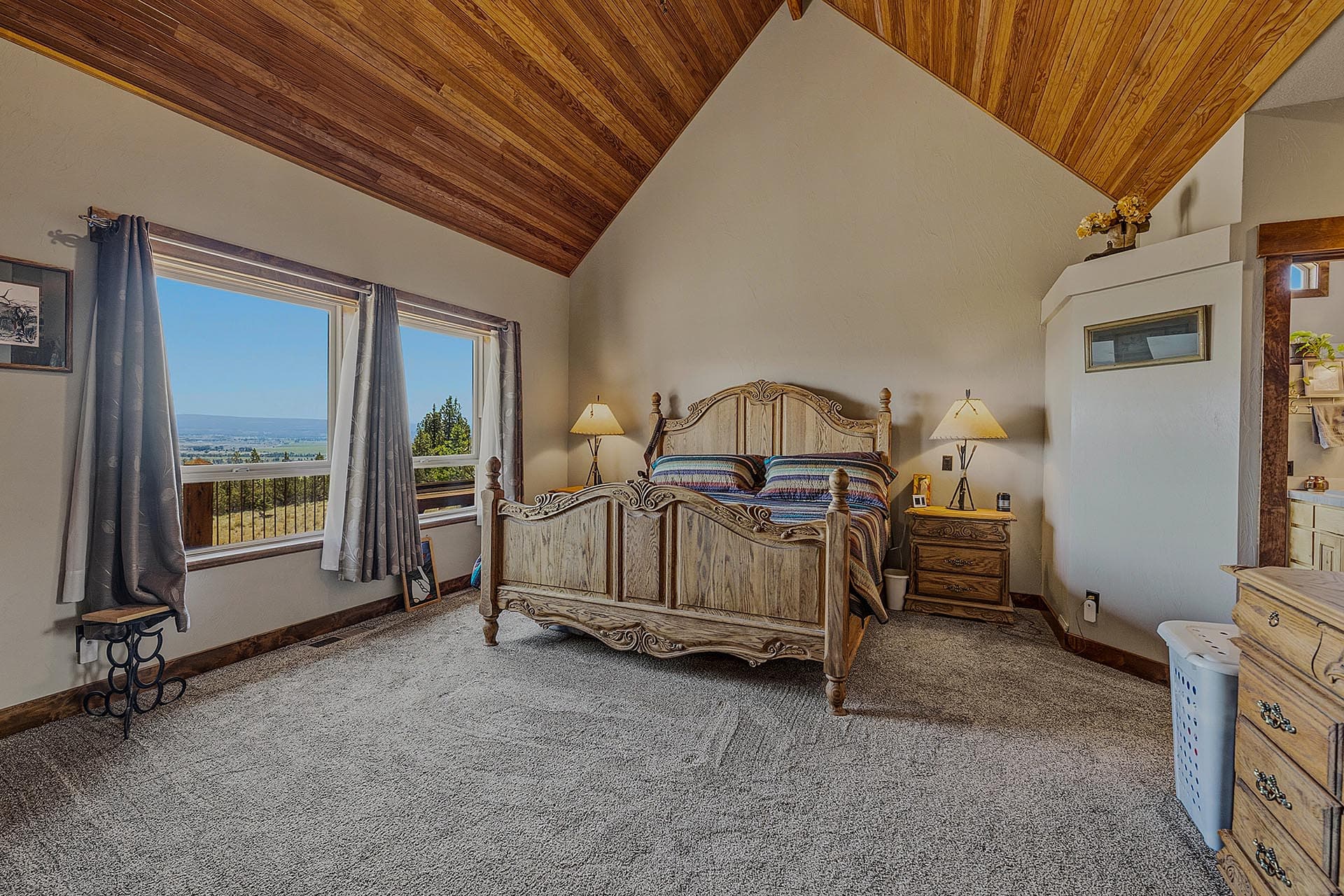 Rustic bedroom with an ornate carved wood bed, two matching nightstands, and a vaulted wood-paneled ceiling, view of landscape through the window.