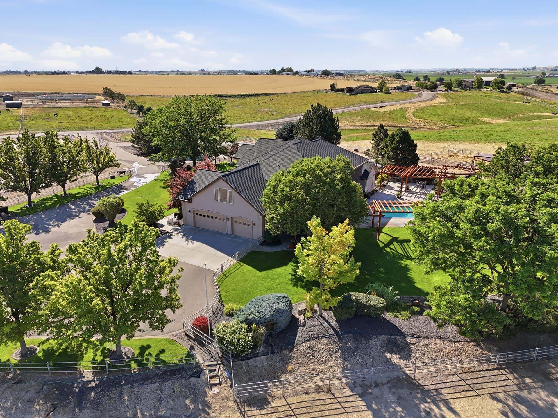 Aerial view of a large backyard with a gray-roofed house, pool under a wooden pergola, and green lawns, with fields beyond Fast farmland in the distance.