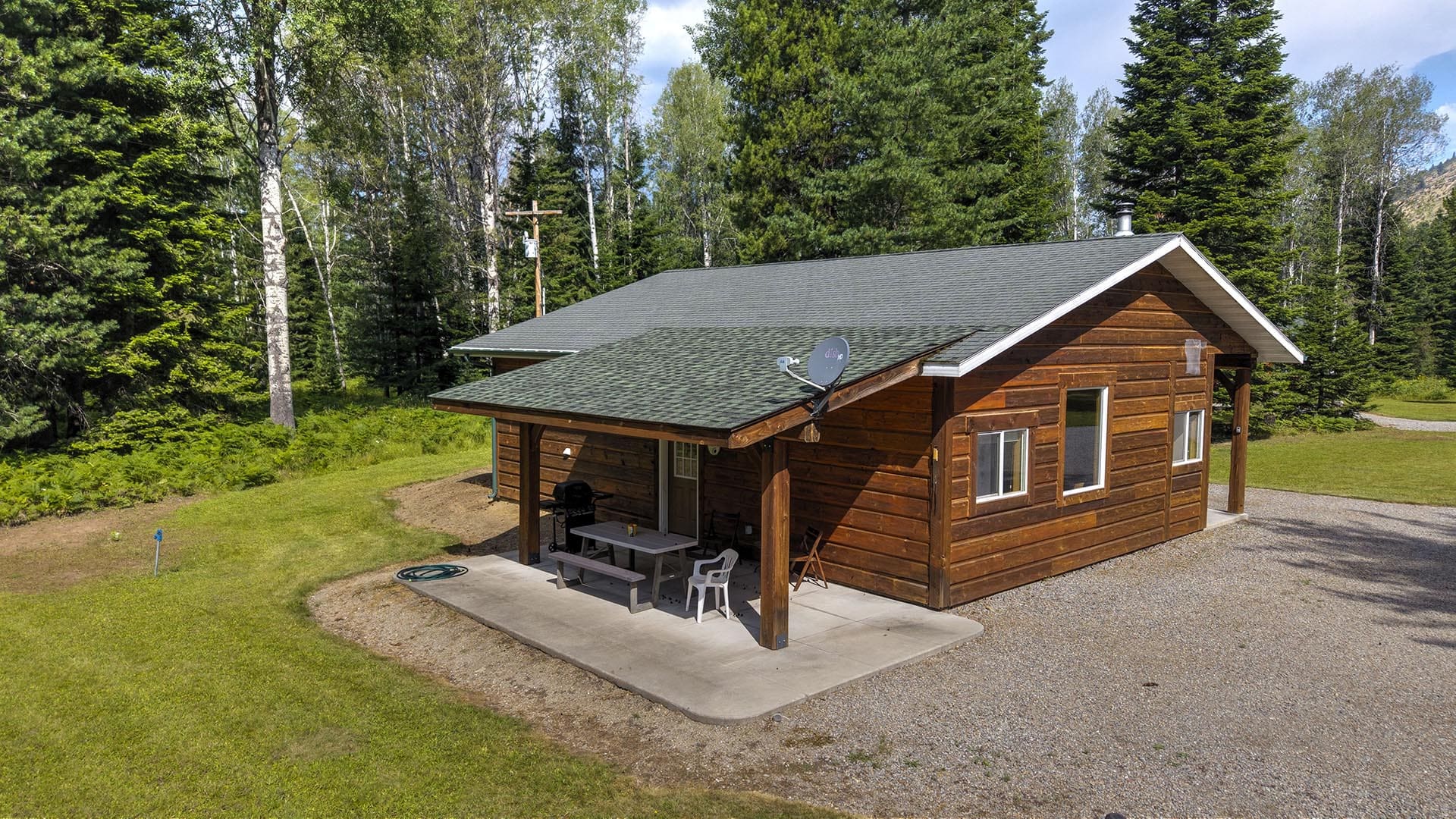 Wooden cabin in a forest clearing with a covered porch, outdoor table and chairs, and a satellite dish on the roof.