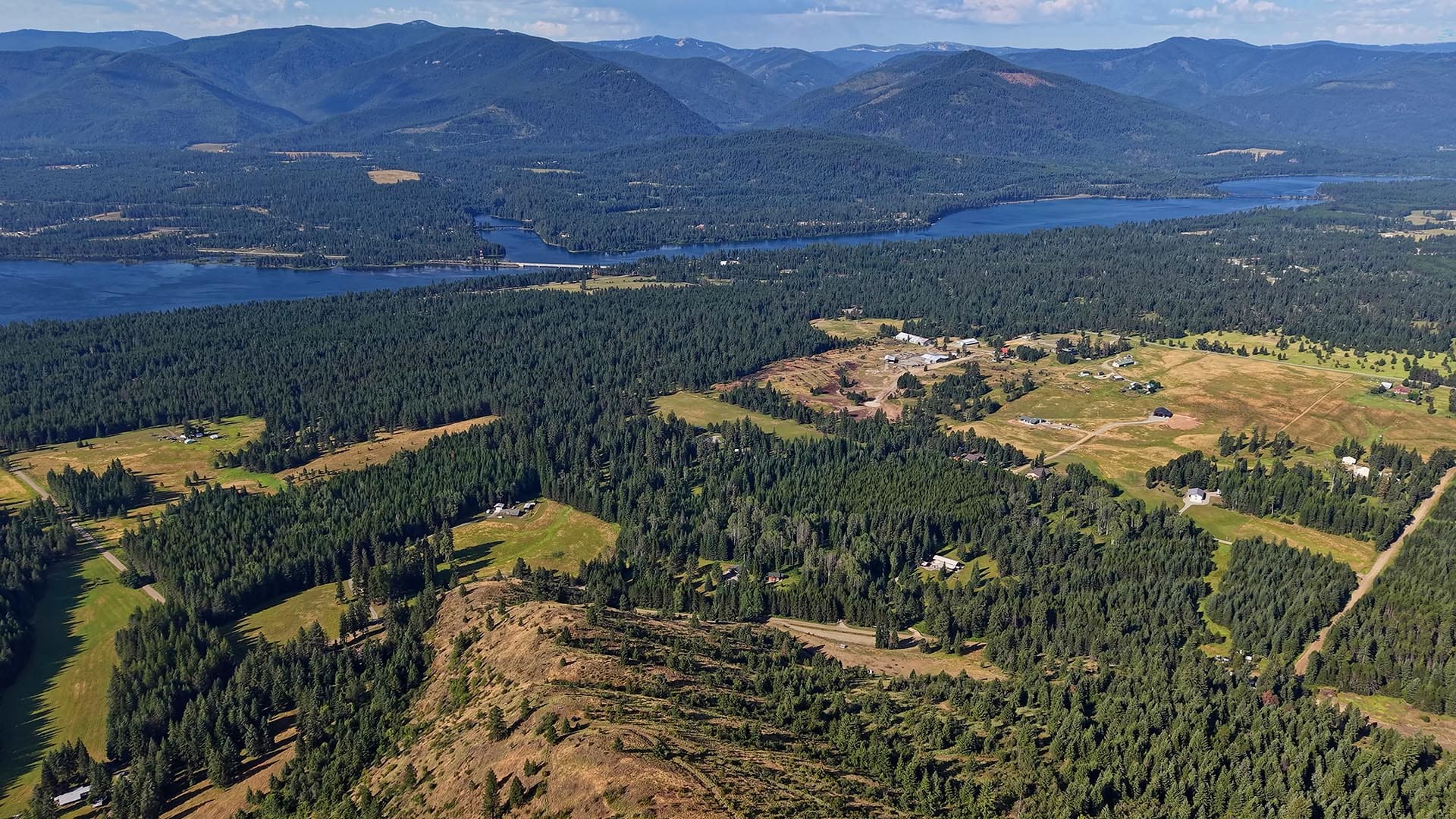Aerial view of a forested valley with a blue lake, distant mountains, and scattered farmhouses and roads.