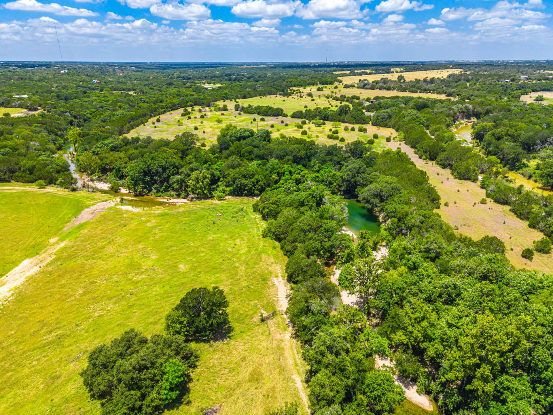 Aerial view of rural countryside with green fields, scattered trees, a winding creek, and a small pond under a blue sky.