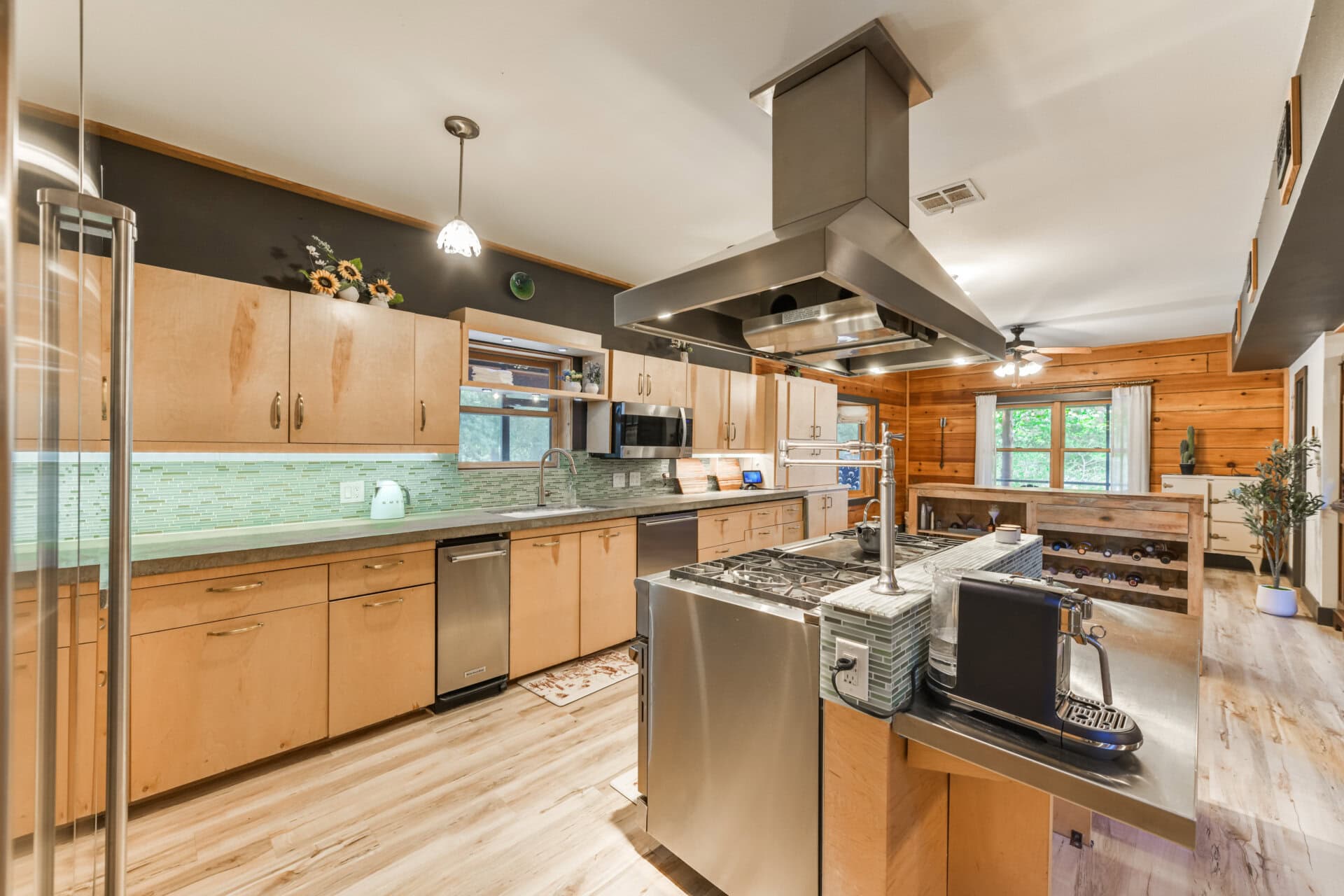Large stainless steel kitchen island with built-in grill and ceiling hood in a wood-and-green-tile kitchen combo.