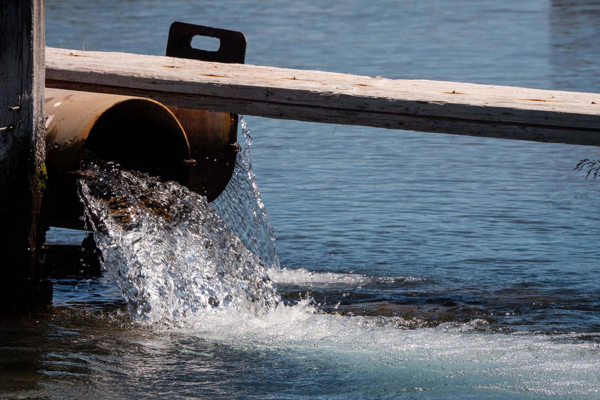 Rusty metal pipe releasing water under a weathered wooden beam into a calm body of water.