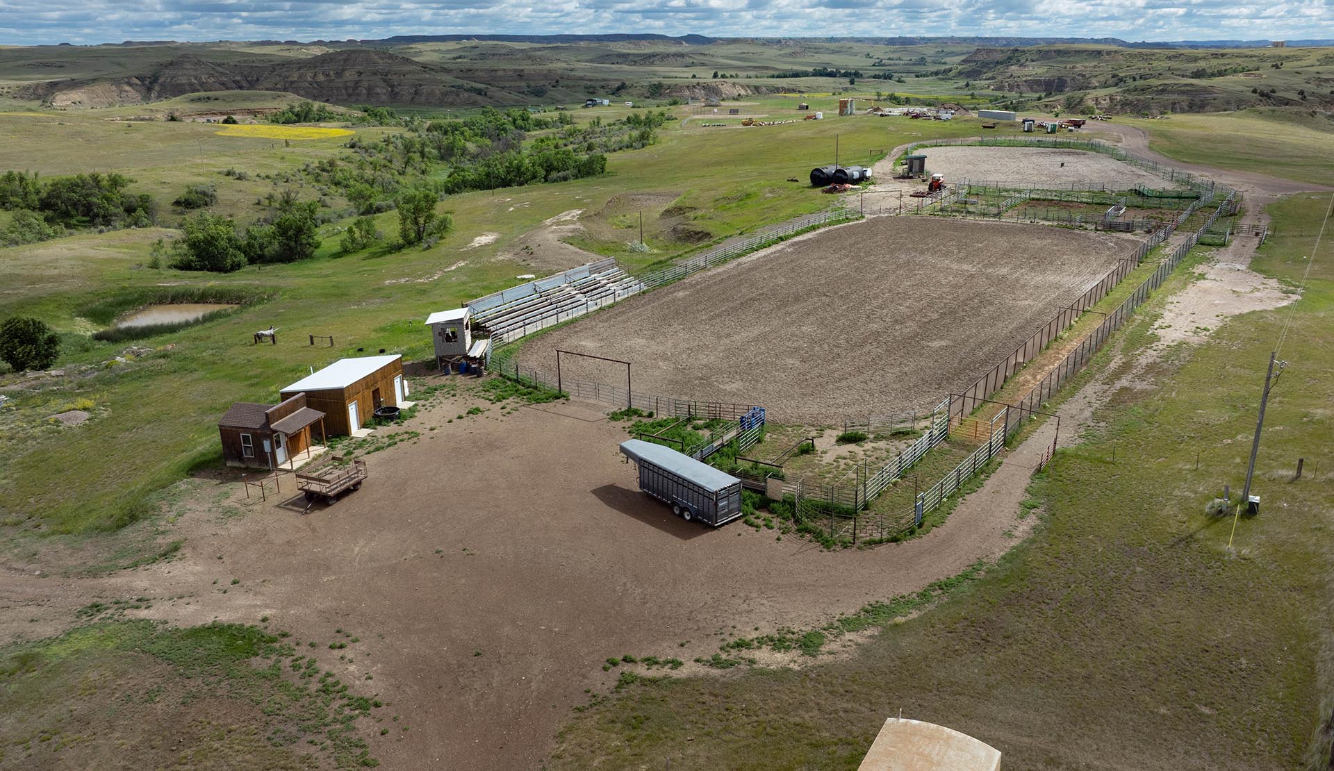 Aerial view of a ranch with a large fenced riding arena, barns, trailers, and open grassy hills.