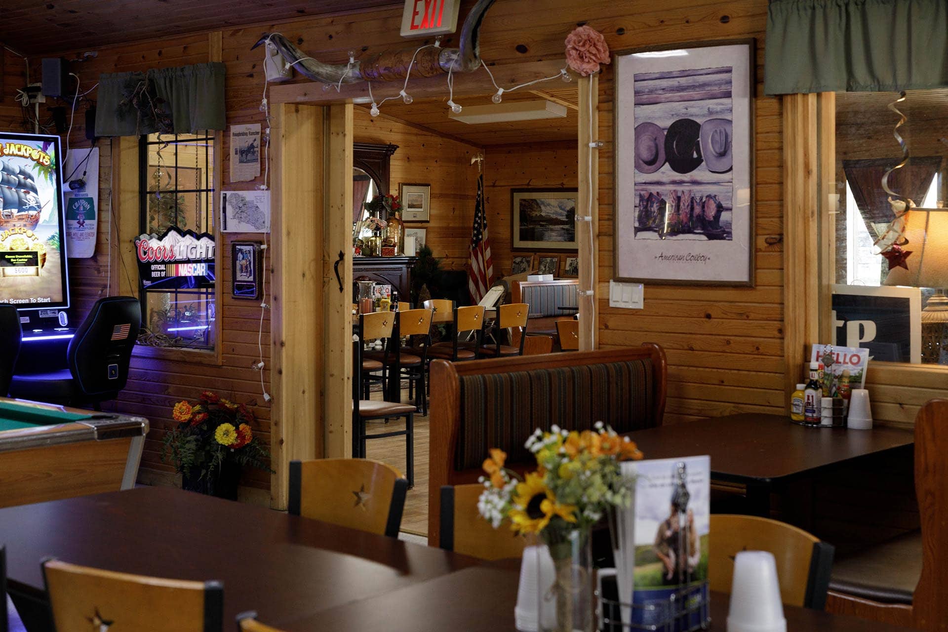 Rustic wood-paneled dining area with flower arrangements on dark tables, framed pictures on the walls, an American flag in the back, and an arcade game visible to the side.