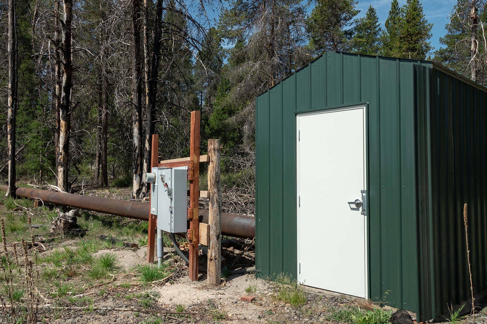 Green metal utility shed with a white door in a forested clearing, with a large brown pipeline and wooden posts nearby.