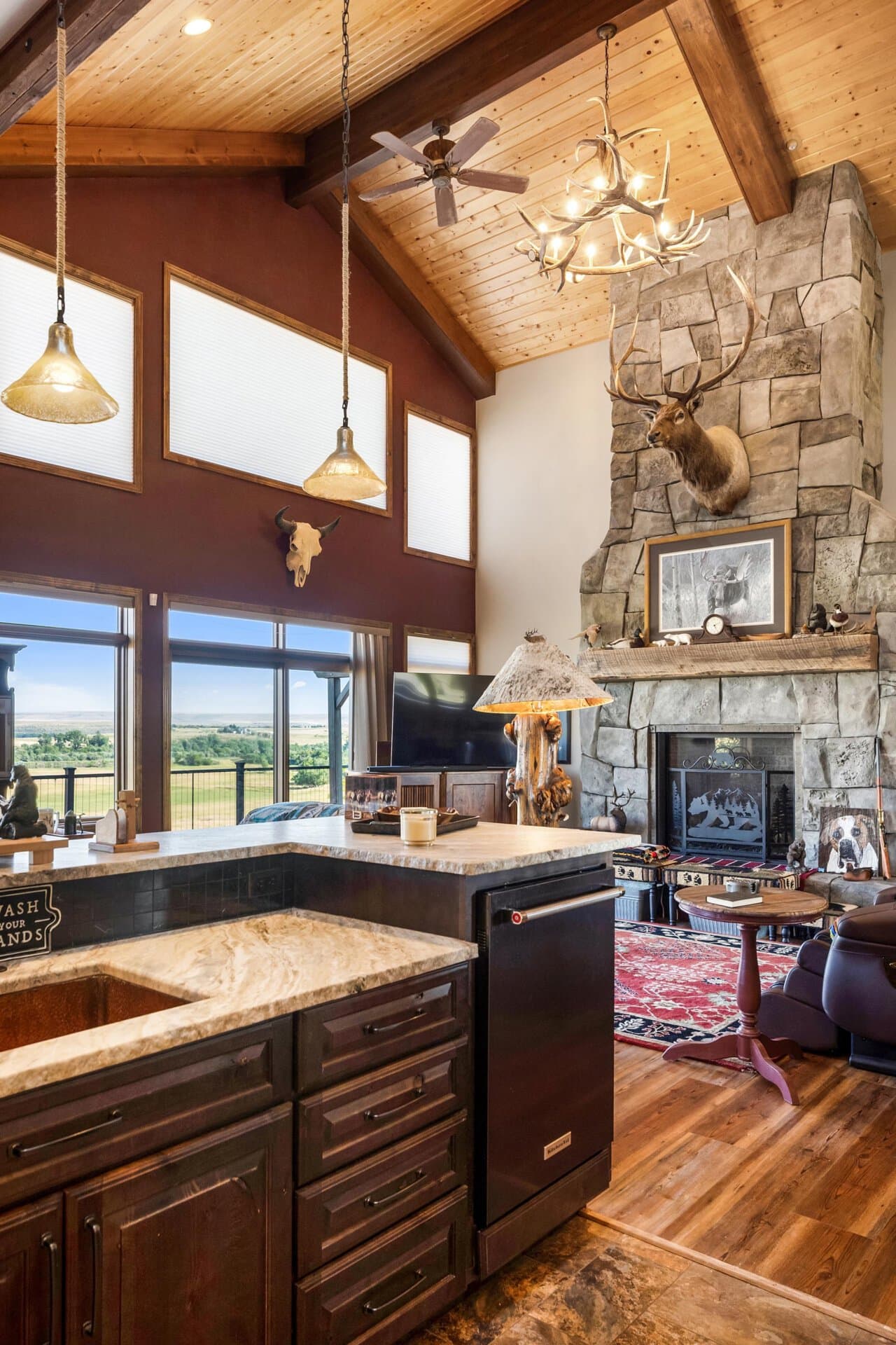 Rustic kitchen with dark wood cabinets, granite island, and a stone fireplace with a mounted elk head.