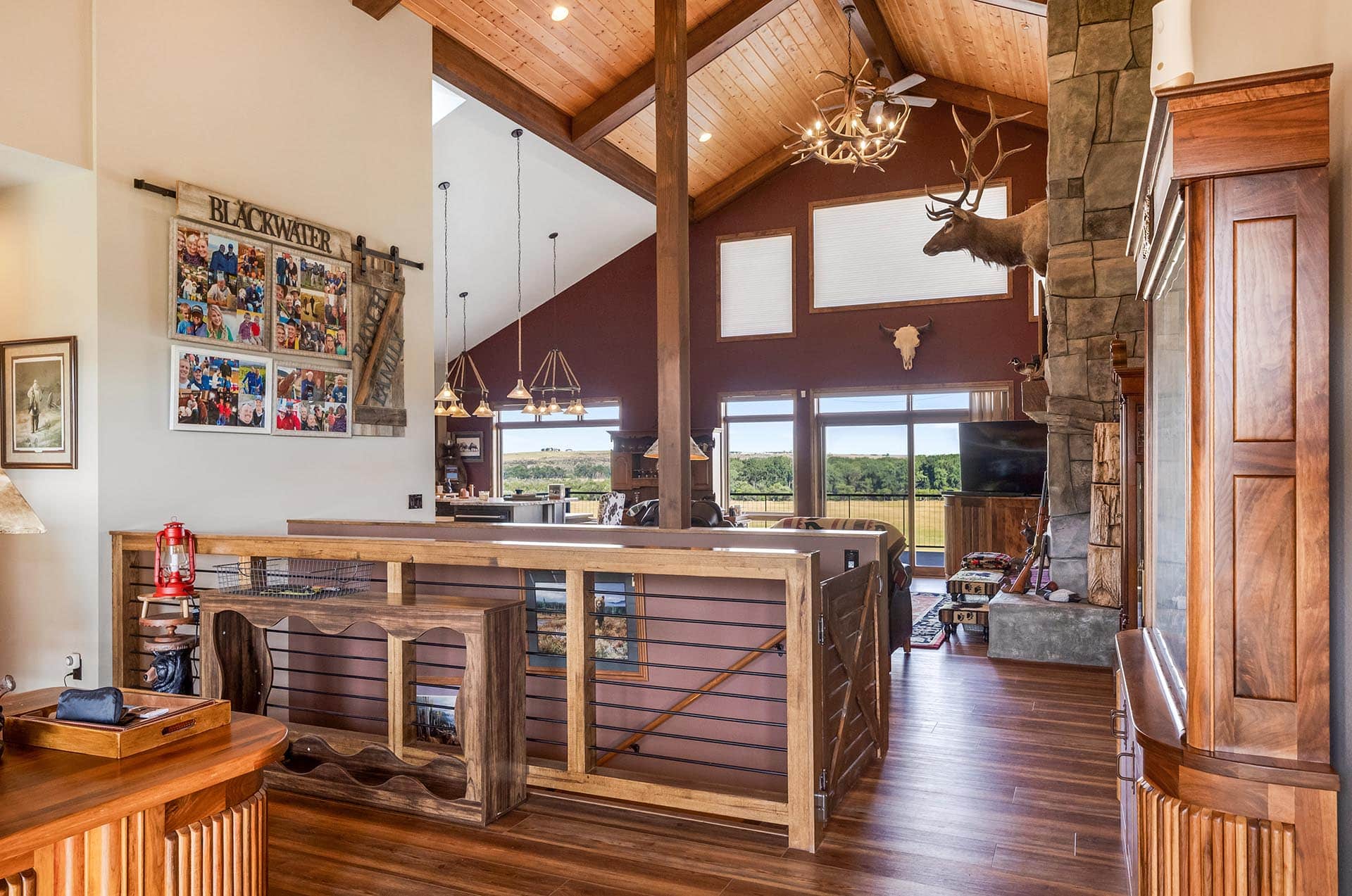 Spacious rustic living area with exposed wood beams, a chandelier of antlers, stone fireplace, and large windows overlooking countryside.