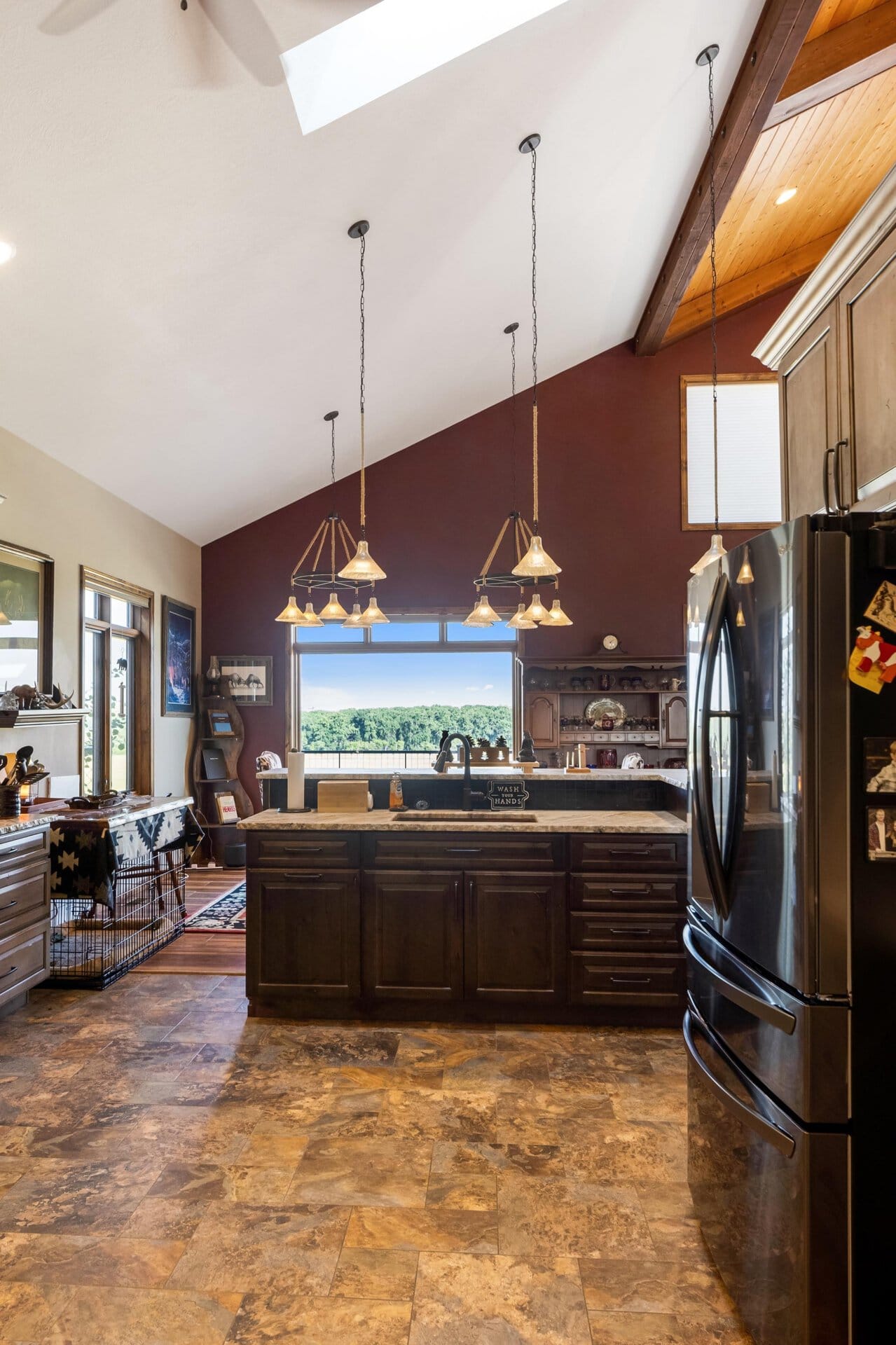 Kitchen with vaulted white ceiling, dark wood island, and hanging pendant lights; stainless steel refrigerator on the right and a window showing greenery outside.
