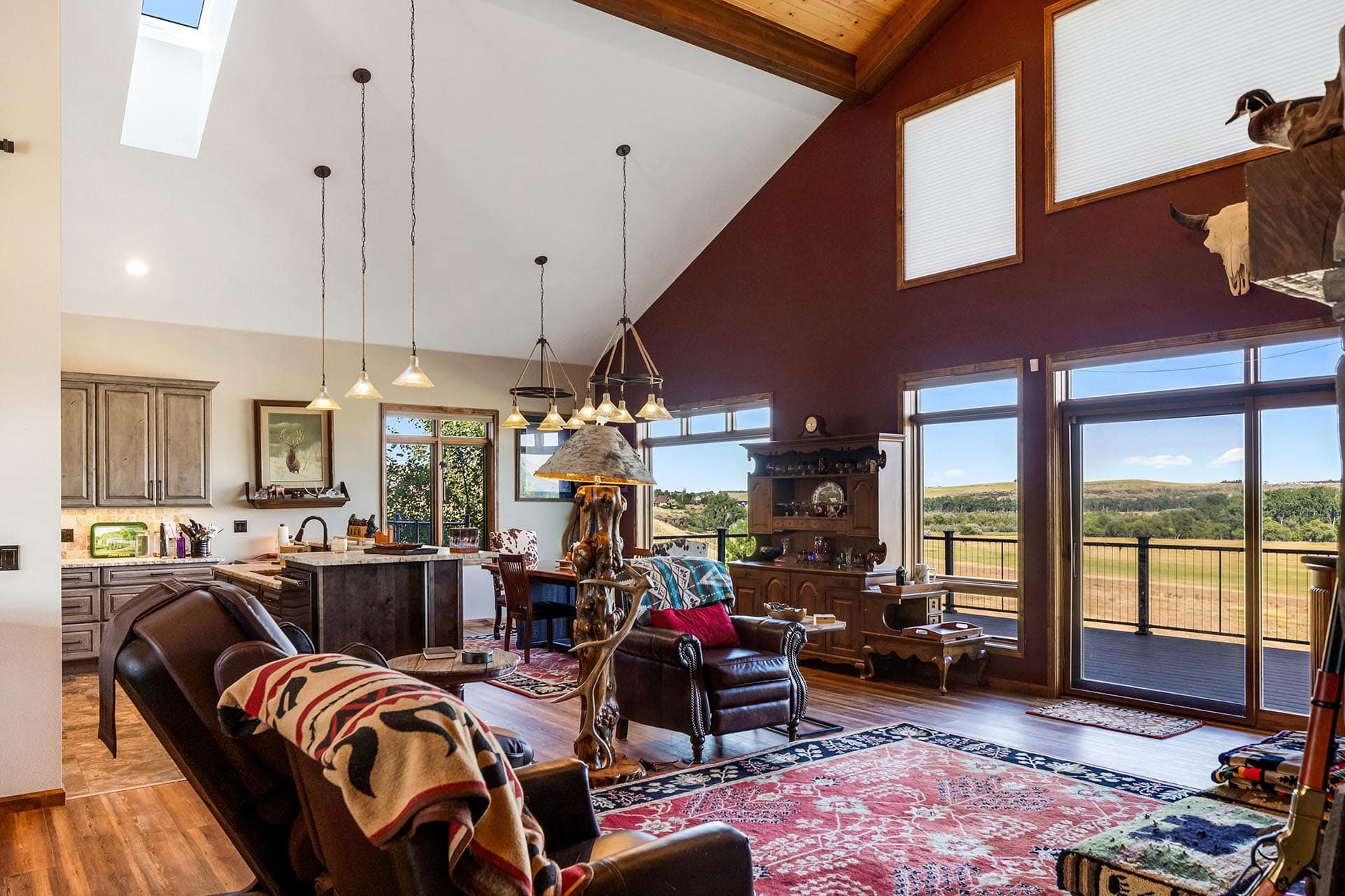Open-concept living area with vaulted white ceiling, rustic wood beams, pendant lights, and large windows showing a rural landscape.