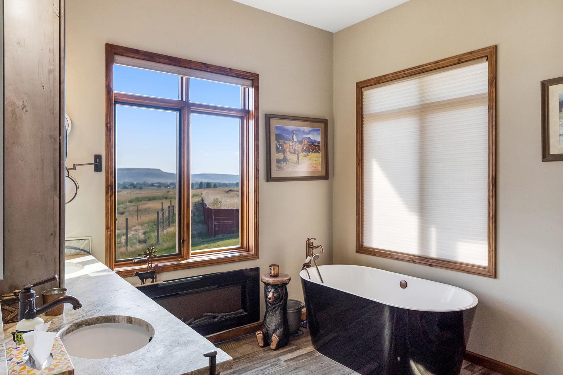 Sunlit bathroom with a black freestanding tub beside a large wood-framed window overlooking fields and hills.