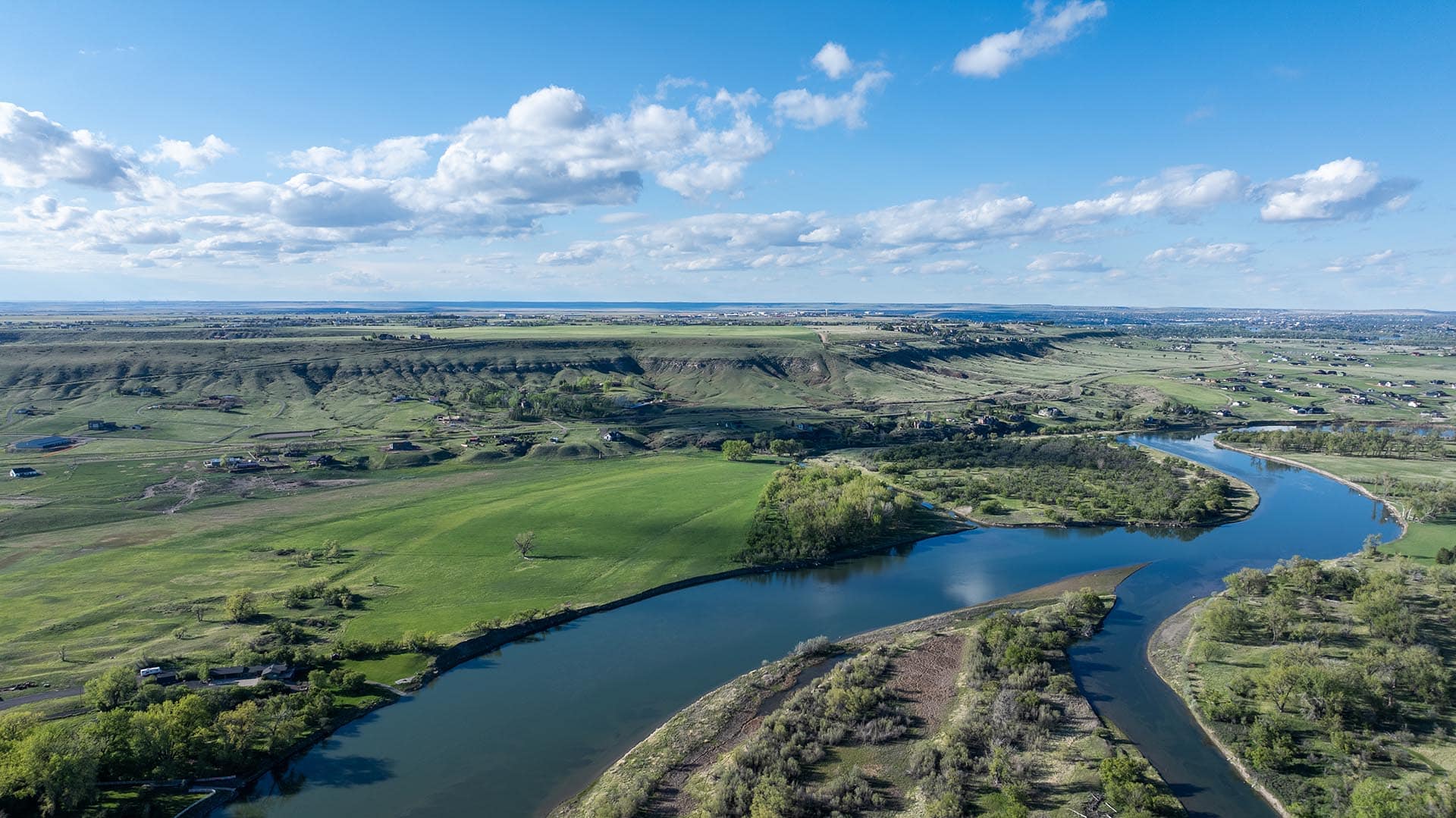 Aerial view of a winding river through green fields and hills with scattered houses under a blue sky.