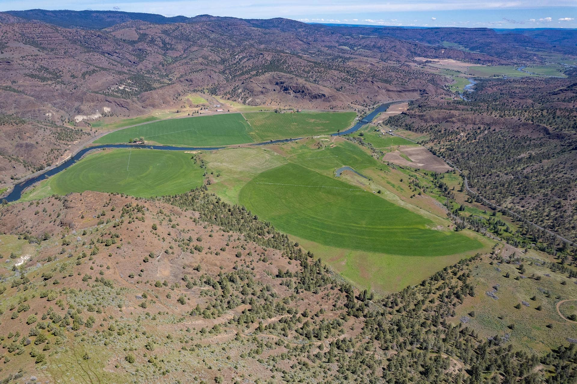 Aerial view of green agricultural fields winding along a dark river with brown hills and sparse trees in the background under a blue sky.