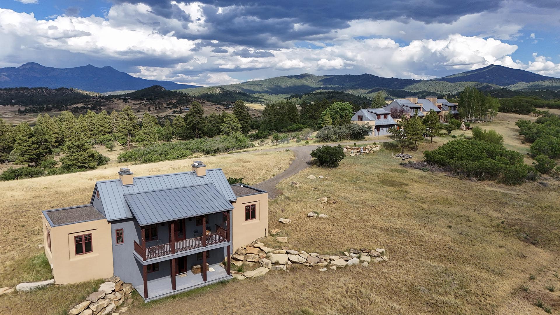 Aerial view of a hillside village with beige houses and metal roofs, winding driveway, and rocky landscape set against pine trees and distant mountains under cloudy skies.