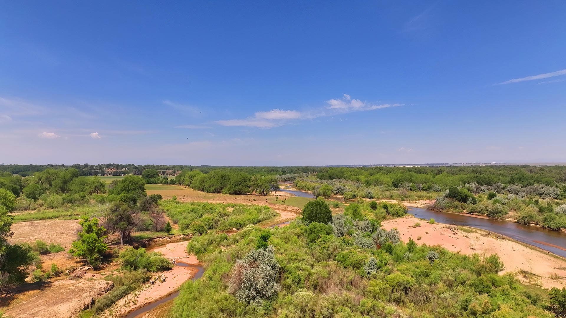 Wide river winding through a green, shrub-filled landscape under a clear blue sky.