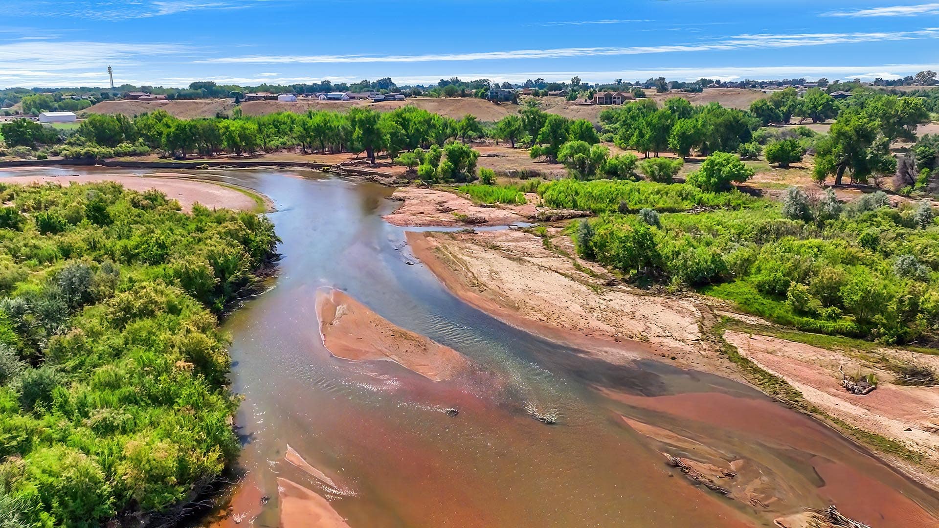 Aerial view of a winding river through a green landscape with lush trees and a sandy shoreline under a bright blue sky.