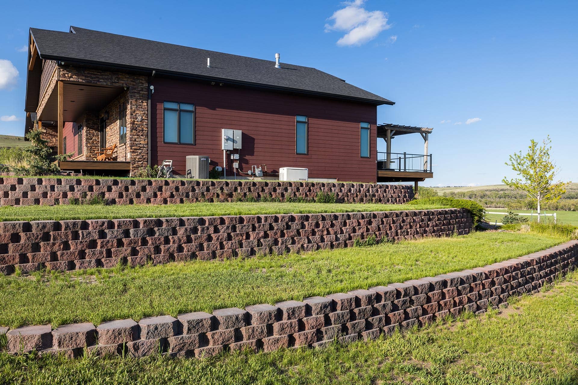 Modern red house with stone facade, raised on a tiered brick retaining wall, under a blue sky.