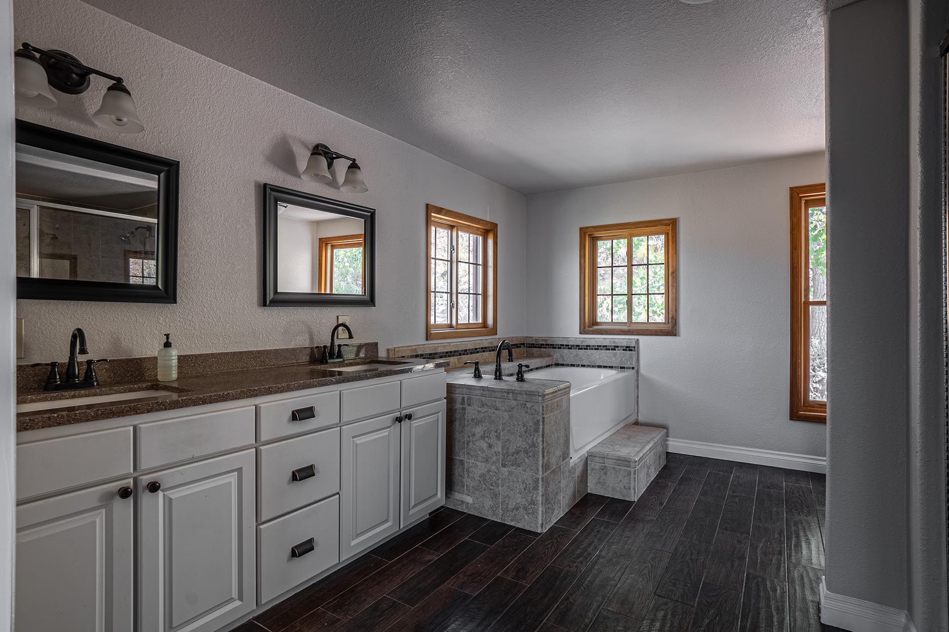 Bathroom with a double vanity, white cabinets, dark granite countertops, and a soaking tub beneath two wood-framed windows.
